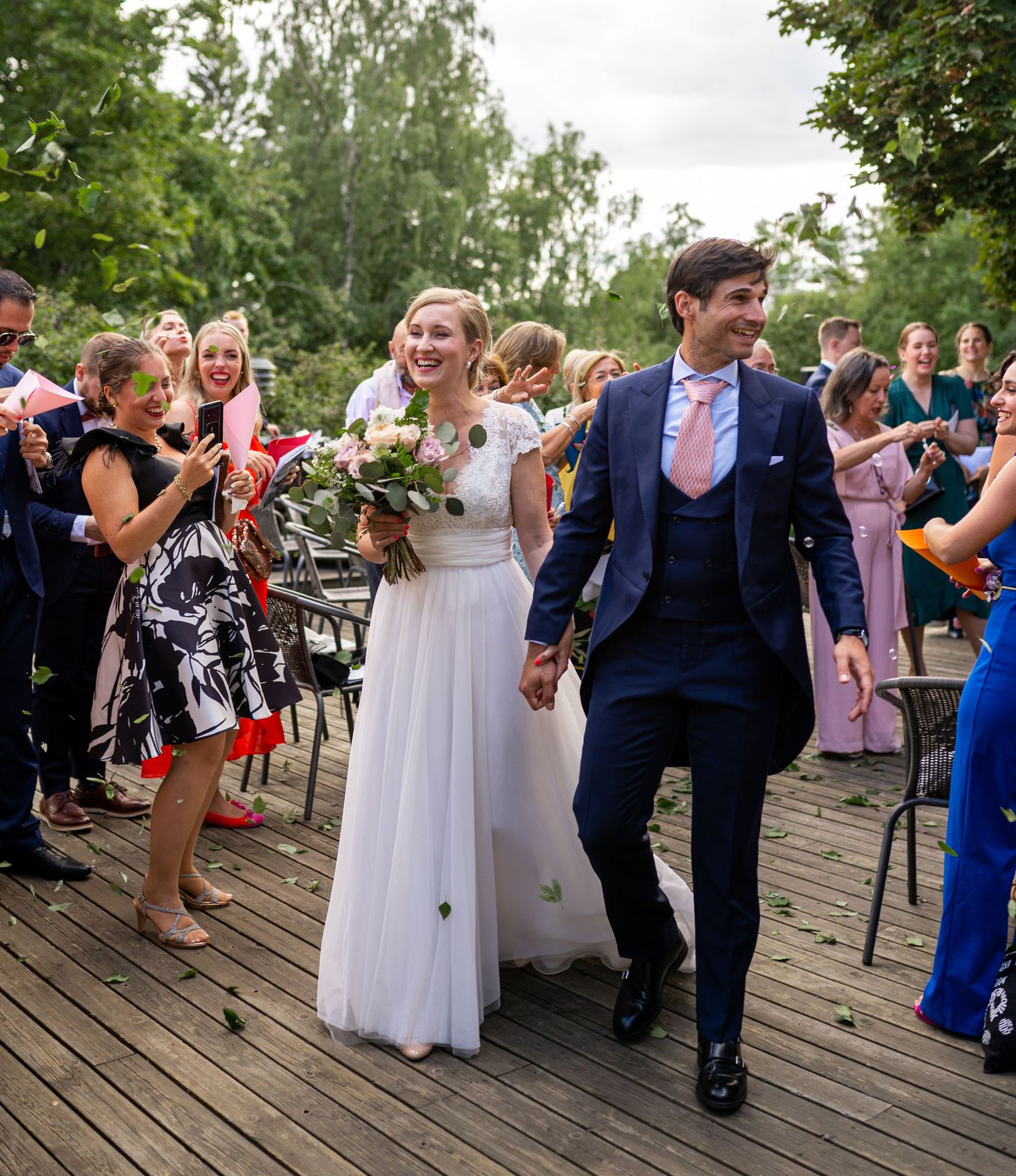 Happy couple exits wedding ceremony as guests cheer and celebrate on a sunlit wooden terrace.