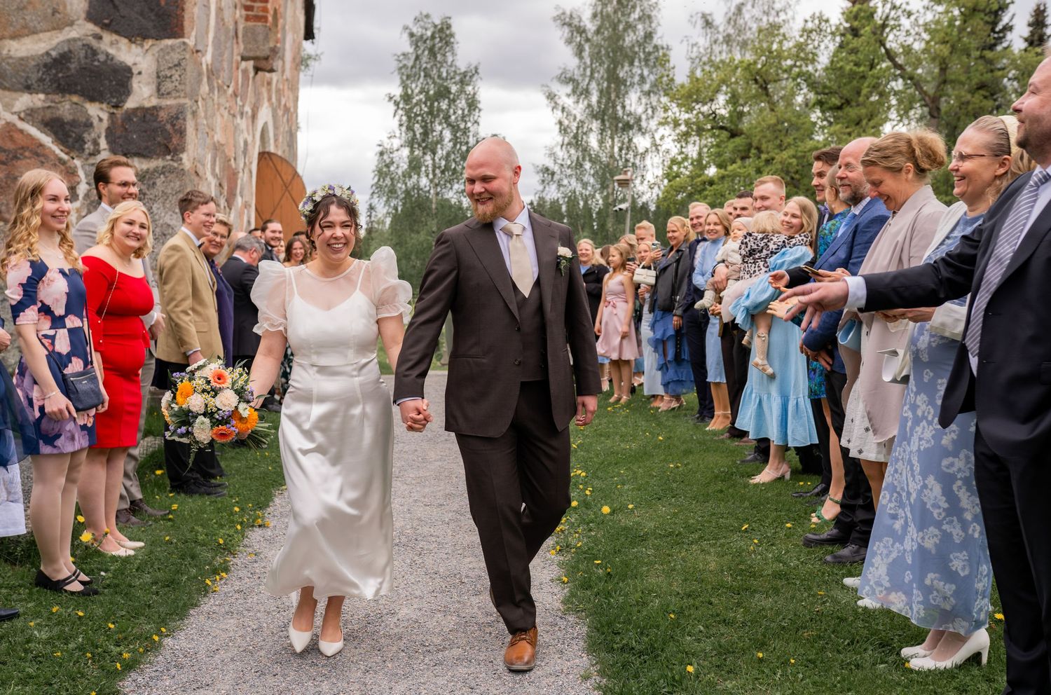Newlyweds walking joyfully through a cheering crowd of guests outside a historic stone church on their wedding day.