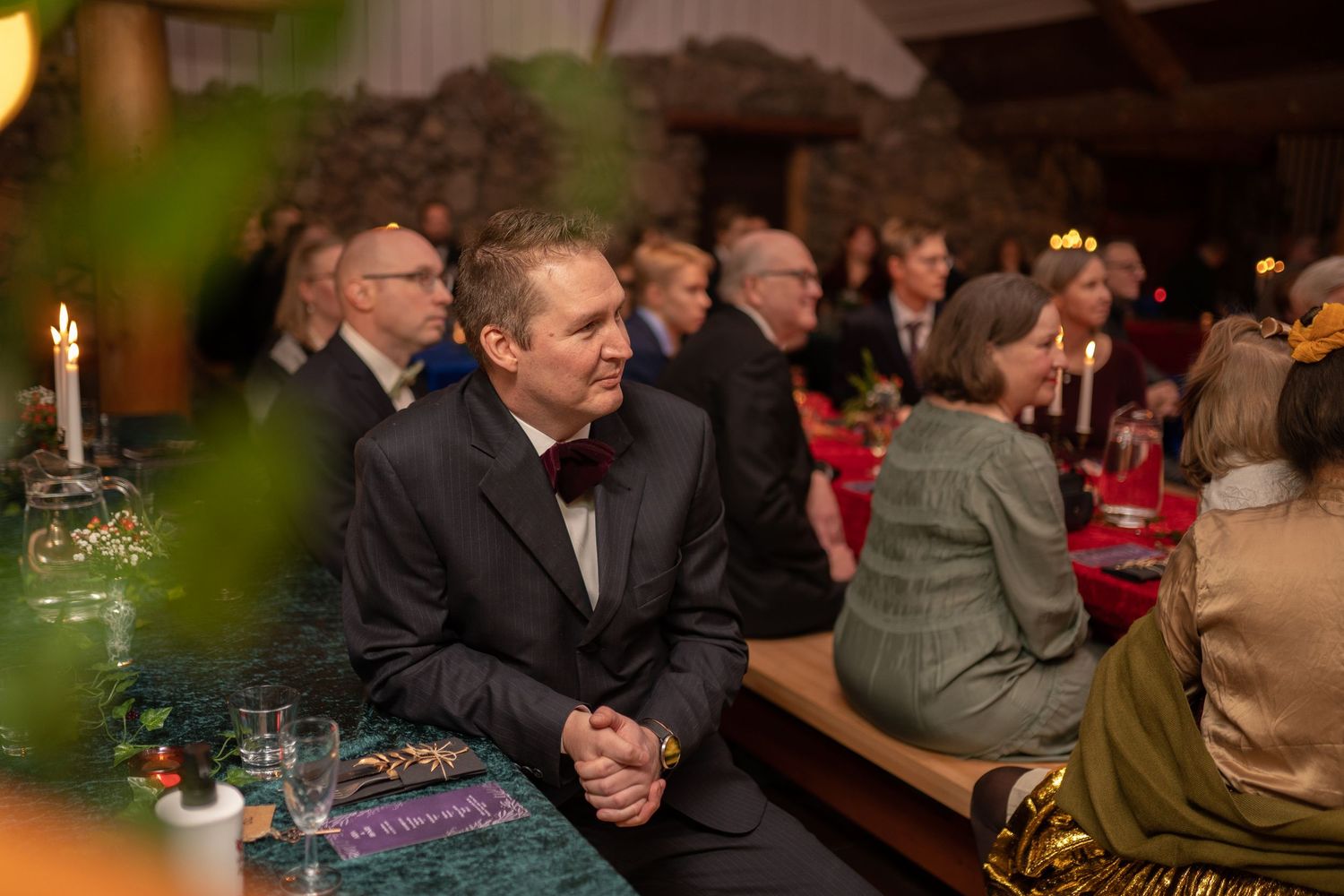 Elegantly dressed guests seated at decorated tables during a formal dinner event in a warmly lit venue.
