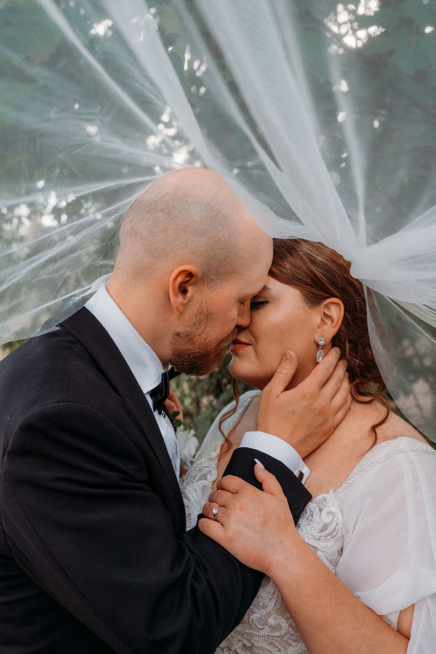 Bald groom kissing bride gently under a billowing white veil outdoors.
