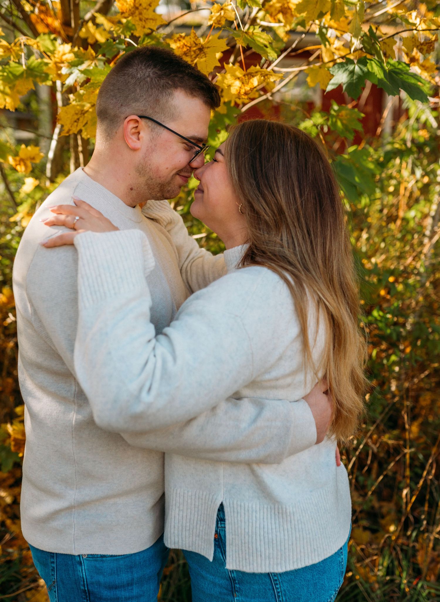 Couple embracing and smiling at each other outdoors among vibrant yellow autumn foliage, wearing matching cream sweaters.