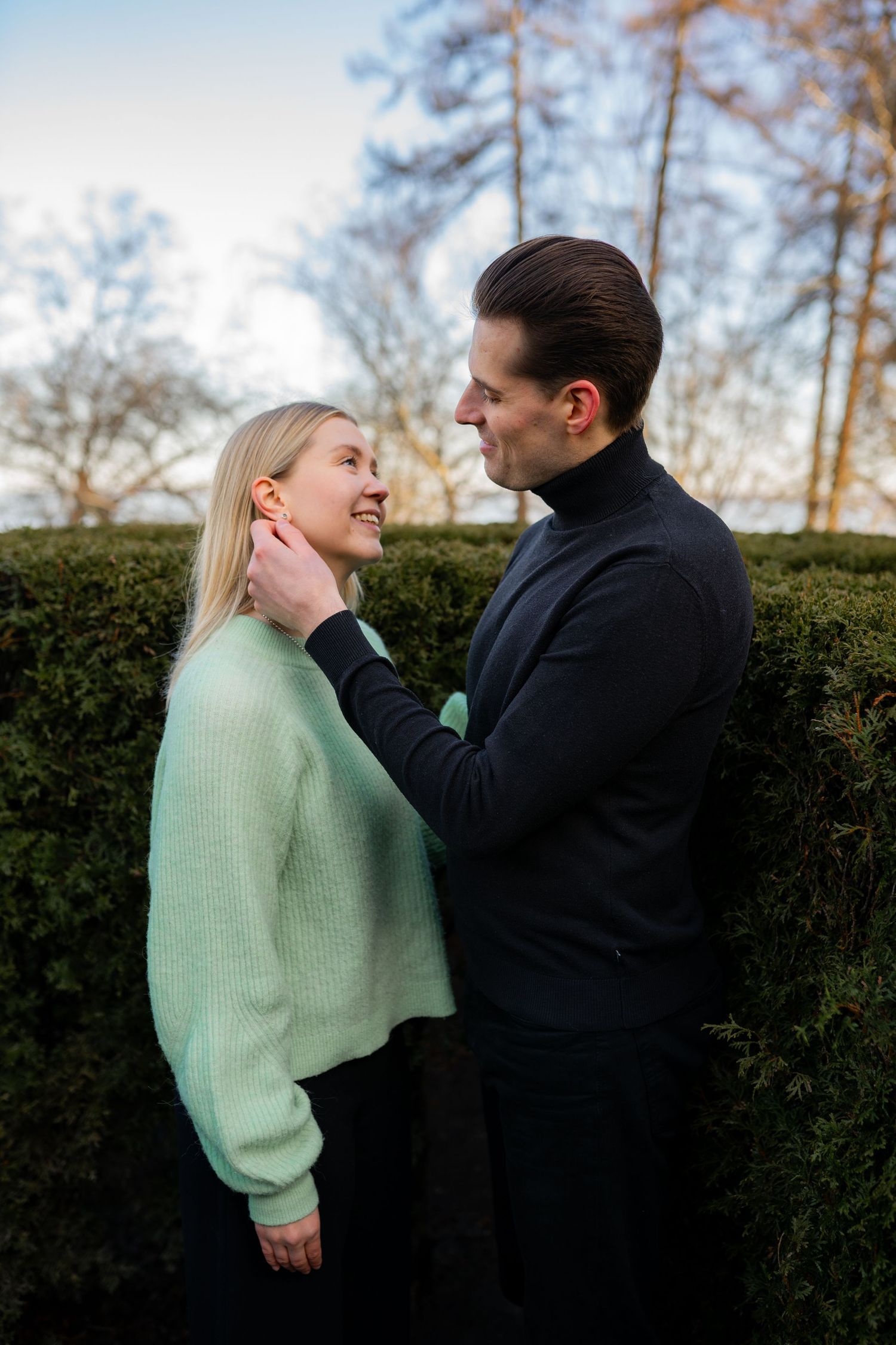 A couple shares a tender moment outdoors, man in black turtleneck touching womans face, she smiles wearing mint green sweater.