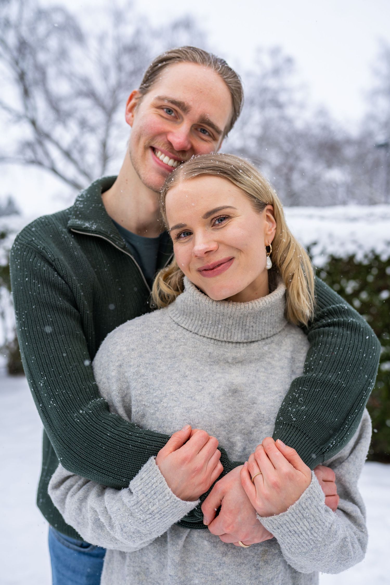 A smiling couple embraces outdoors in a snowy winter setting, wearing cozy gray and green sweaters.
