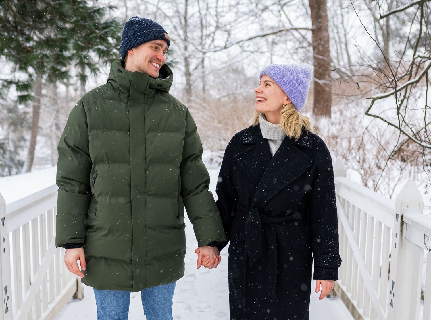Couple holding hands on snowy bridge in winter coats and hats, smiling at each other in a snowy park.