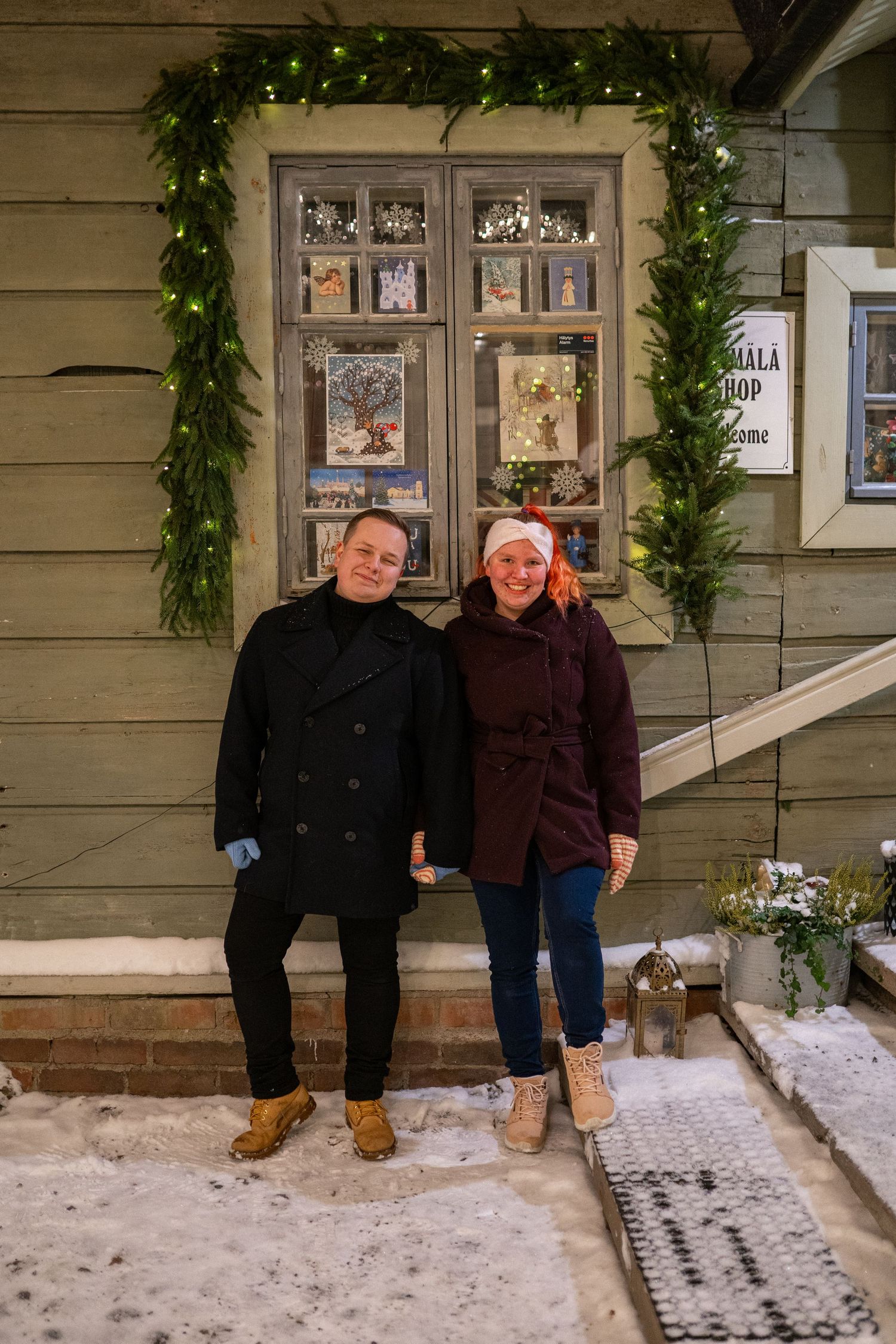 Couple smiling in front of a Christmas-decorated wooden shop entrance with garland lights and snow on the ground.