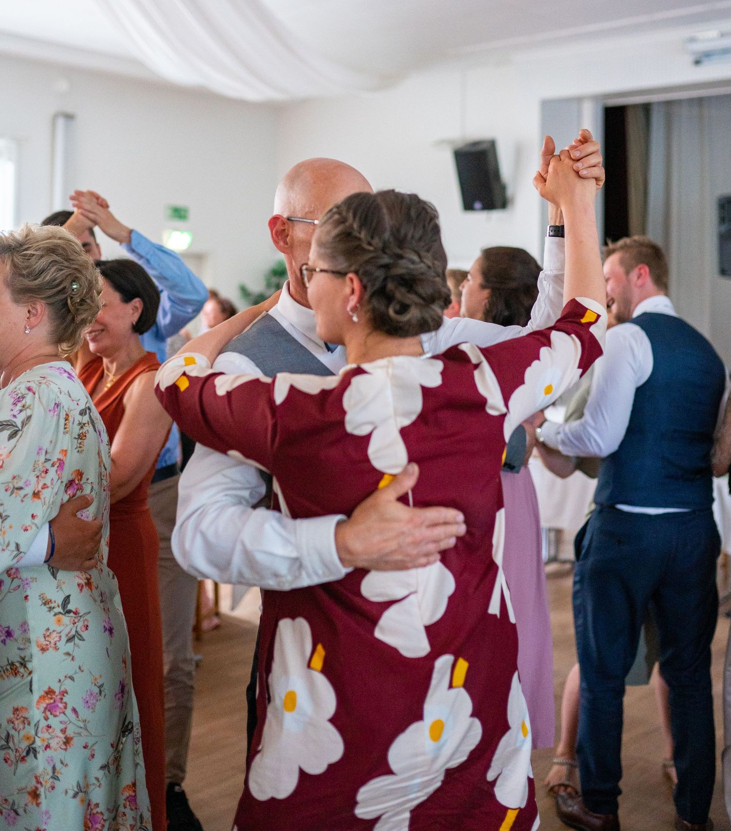 Wedding guests dancing together in a lively celebration, woman in burgundy floral dress prominent in foreground.