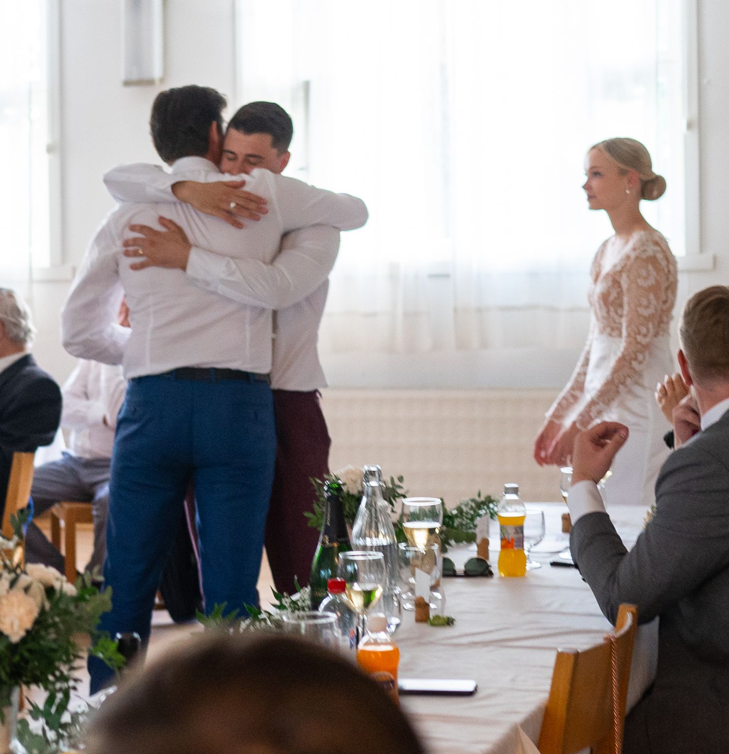 Emotional moment as two men embrace at a wedding reception while the bride in white lace looks on.