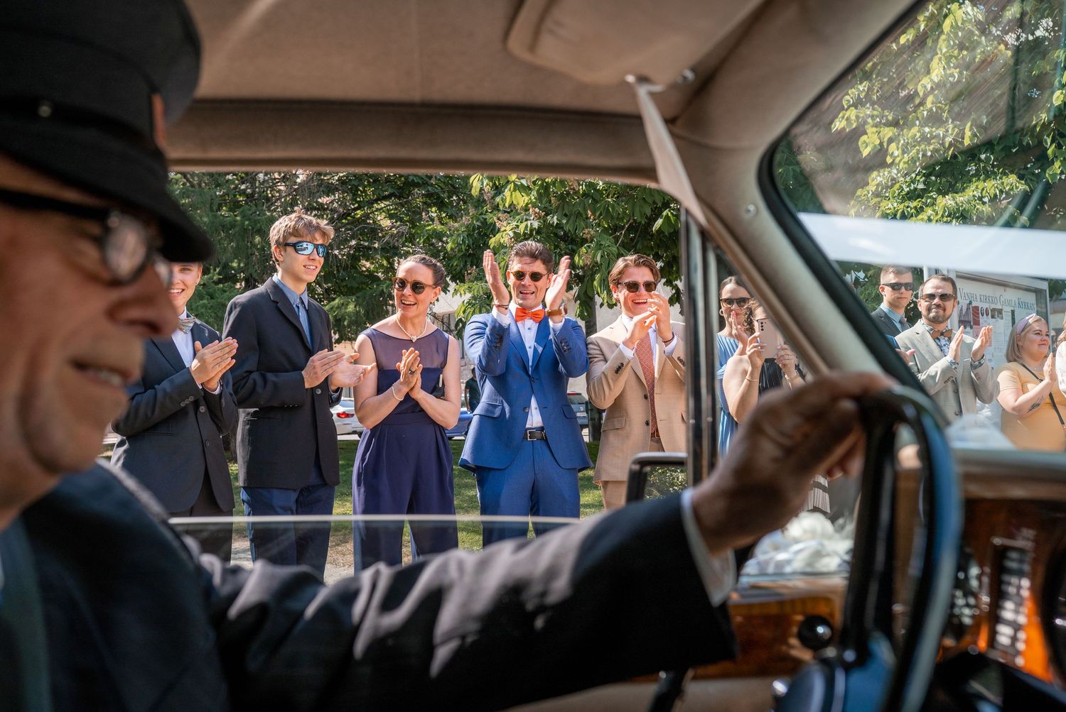Wedding guests applaud as viewed through vintage car windshield mirror on sunny celebration day.