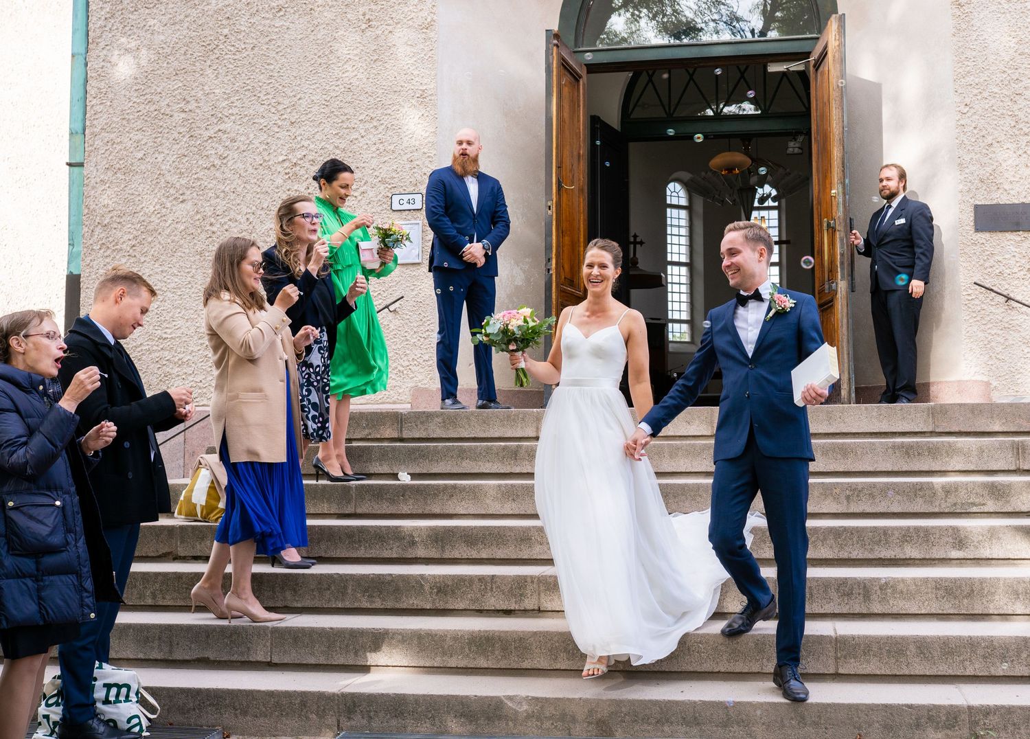Smiling bride and groom walk down church steps holding hands with guests.