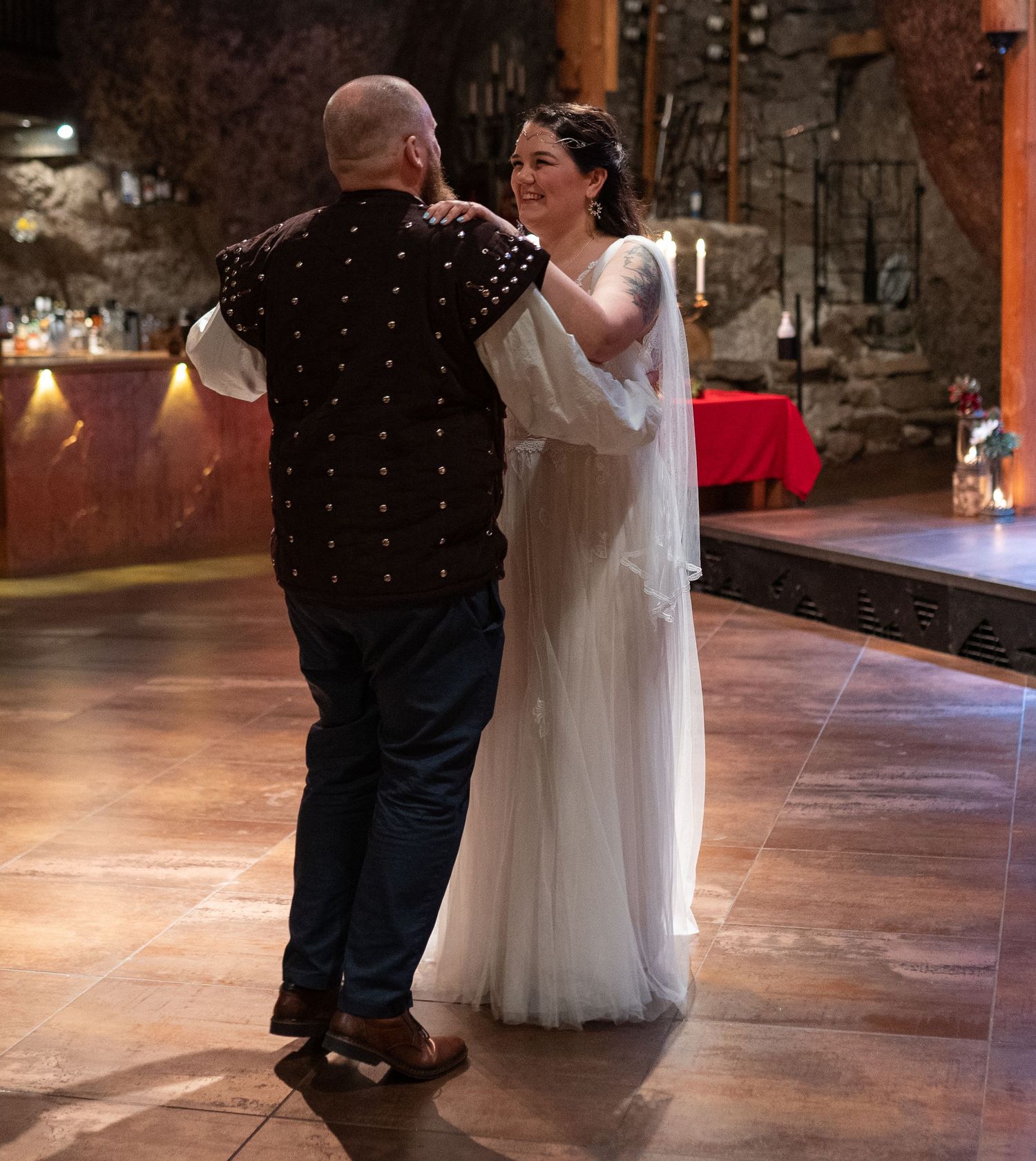 Couple sharing first dance in medieval hall, bride in white gown, groom in dark vest, romantic wedding reception.