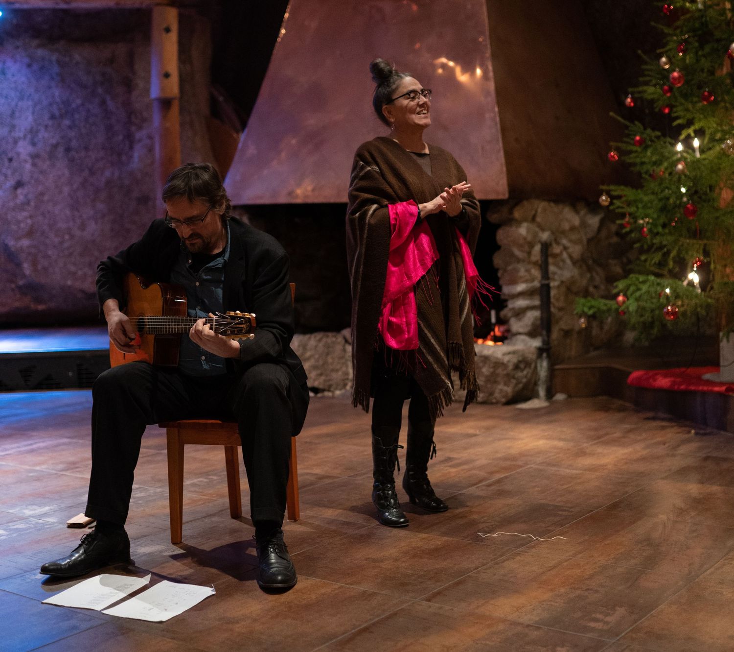 A guitarist and a singer perform indoors near a Christmas tree and fireplace in a festive holiday setting.
