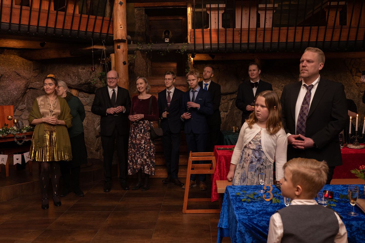 Formal gathering of adults and children in a rustic stone-walled venue with festive decorations and elegant table settings.