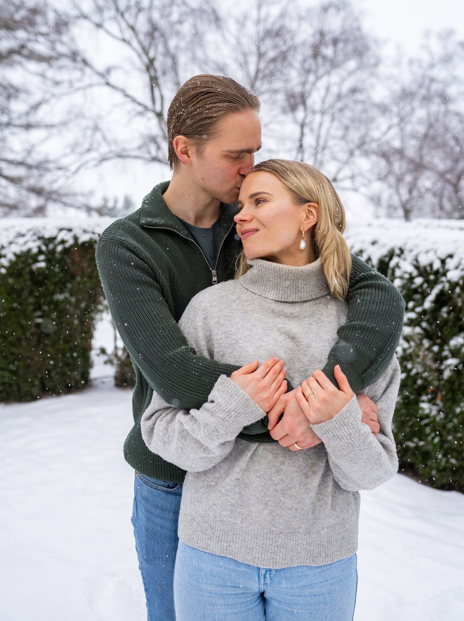 Couple embracing in snowy winter garden, man in green sweater kissing woman in gray turtleneck.