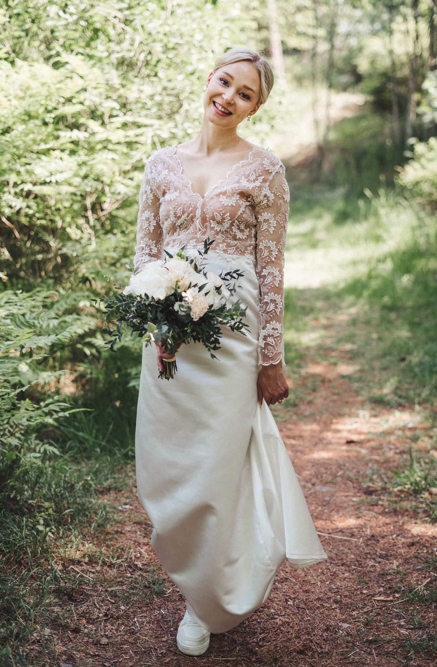 Bride in lace top and satin skirt walking forest path holding white floral bouquet.