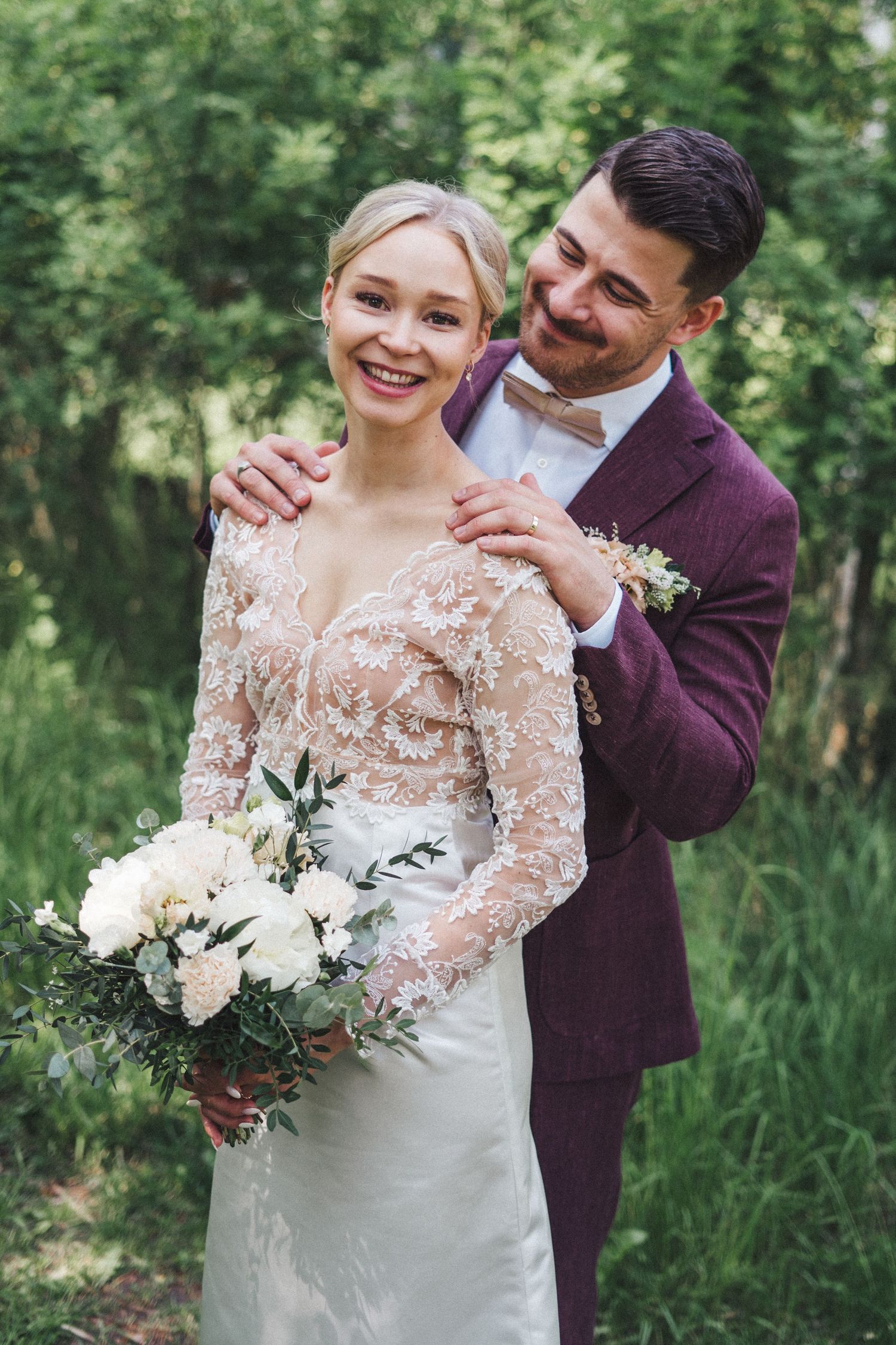 Groom in burgundy suit embracing smiling bride from behind in green garden, bride holding white floral bouquet.
