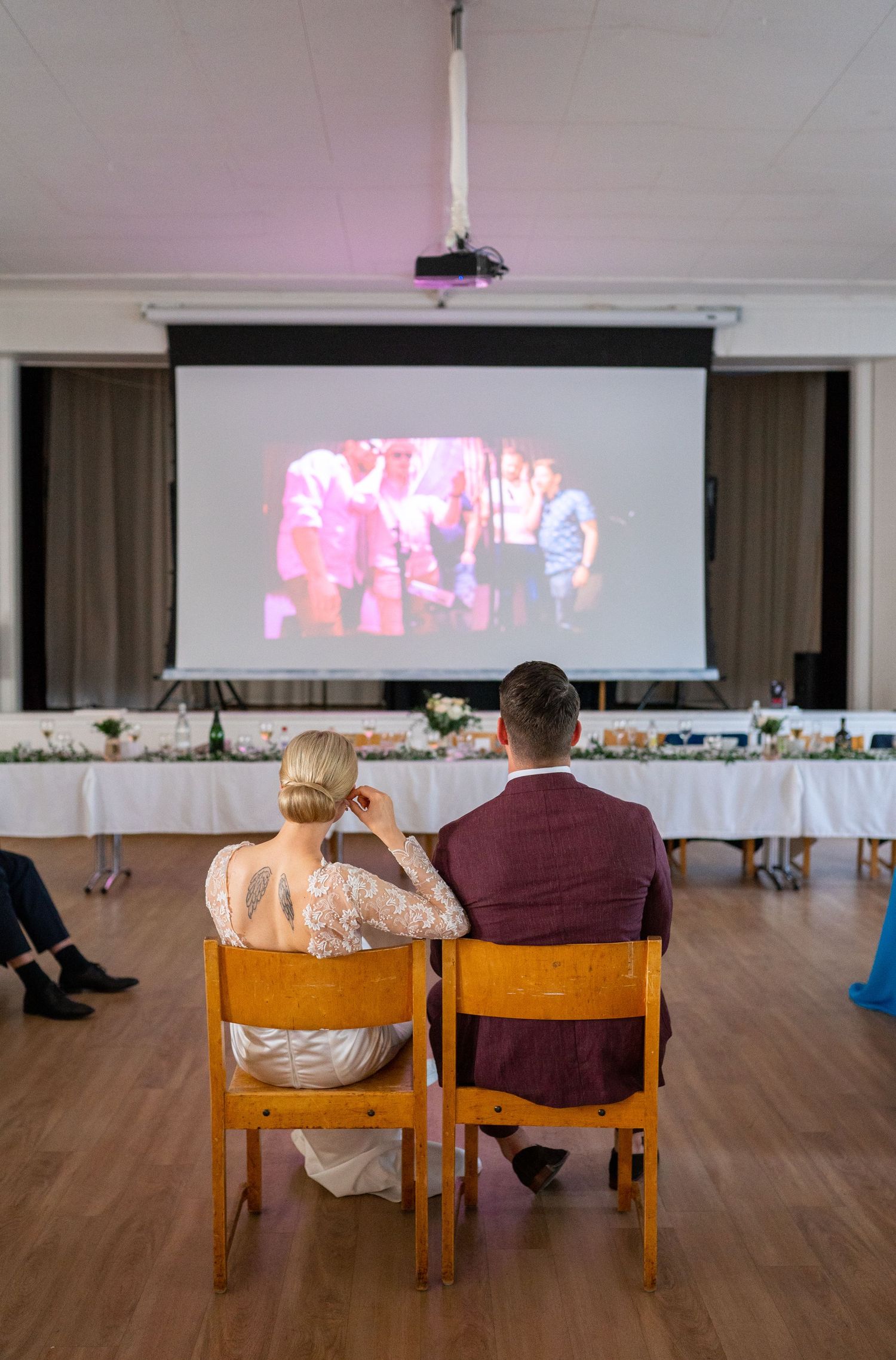Bride and groom seated watching a video presentation on a large screen at their wedding reception.