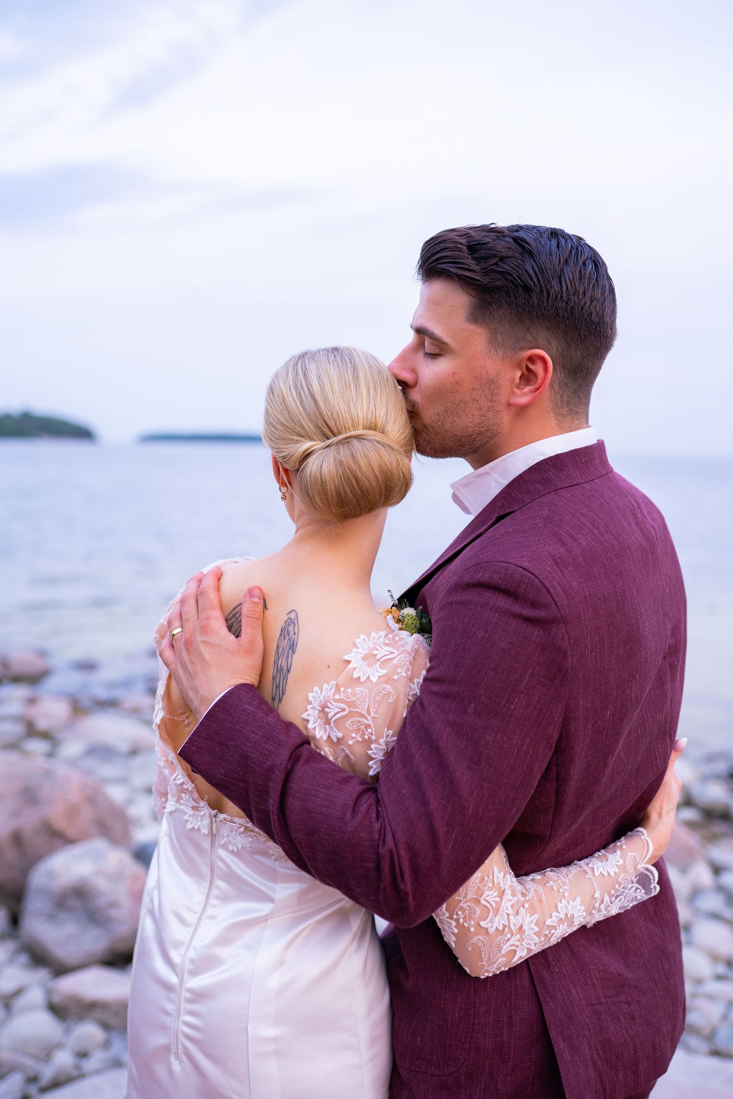 Groom in burgundy suit kissing bride on cheek from behind while embracing on a scenic rocky coastline.