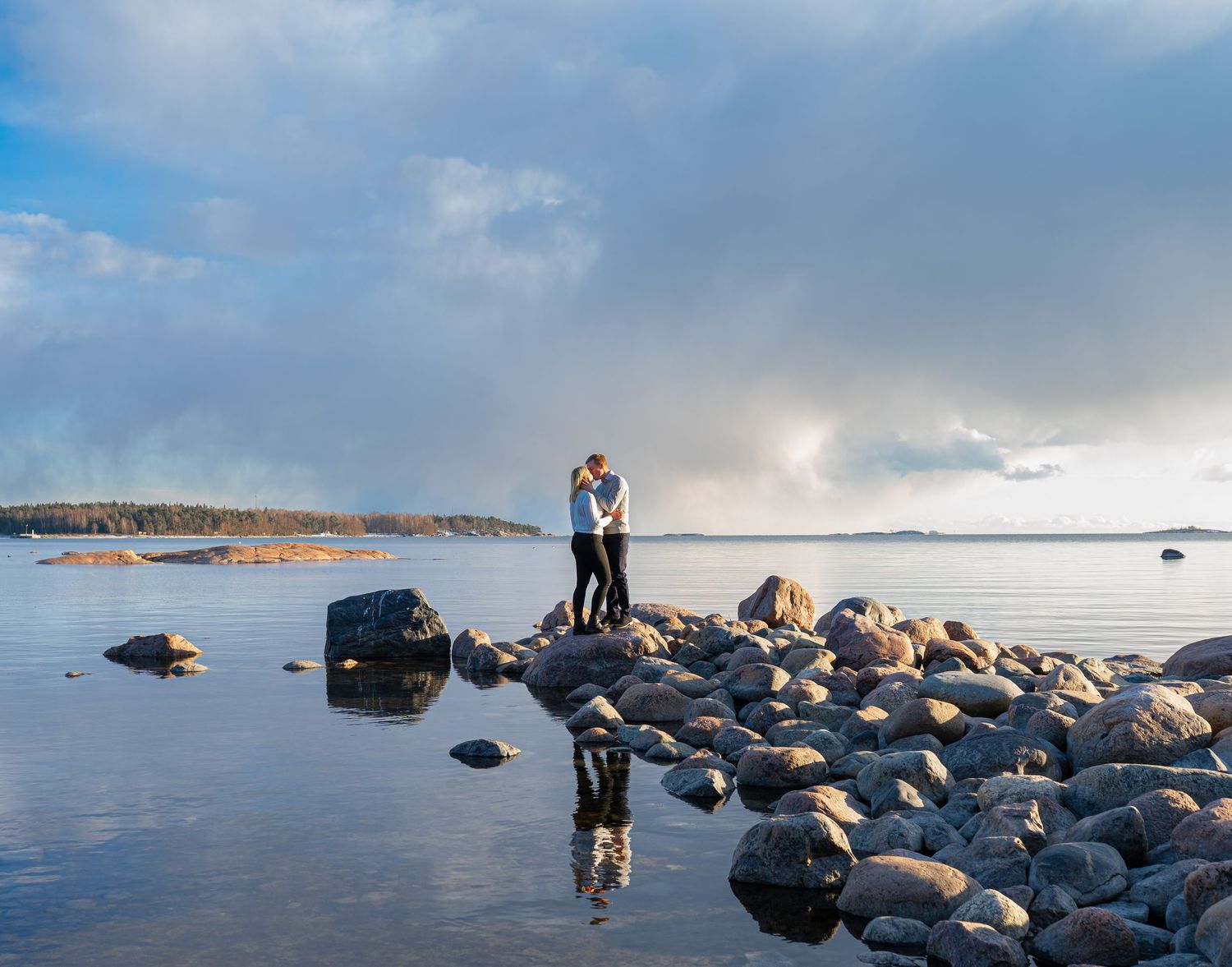 Couple embracing on rocky shore reflected in calm water under dramatic sky.