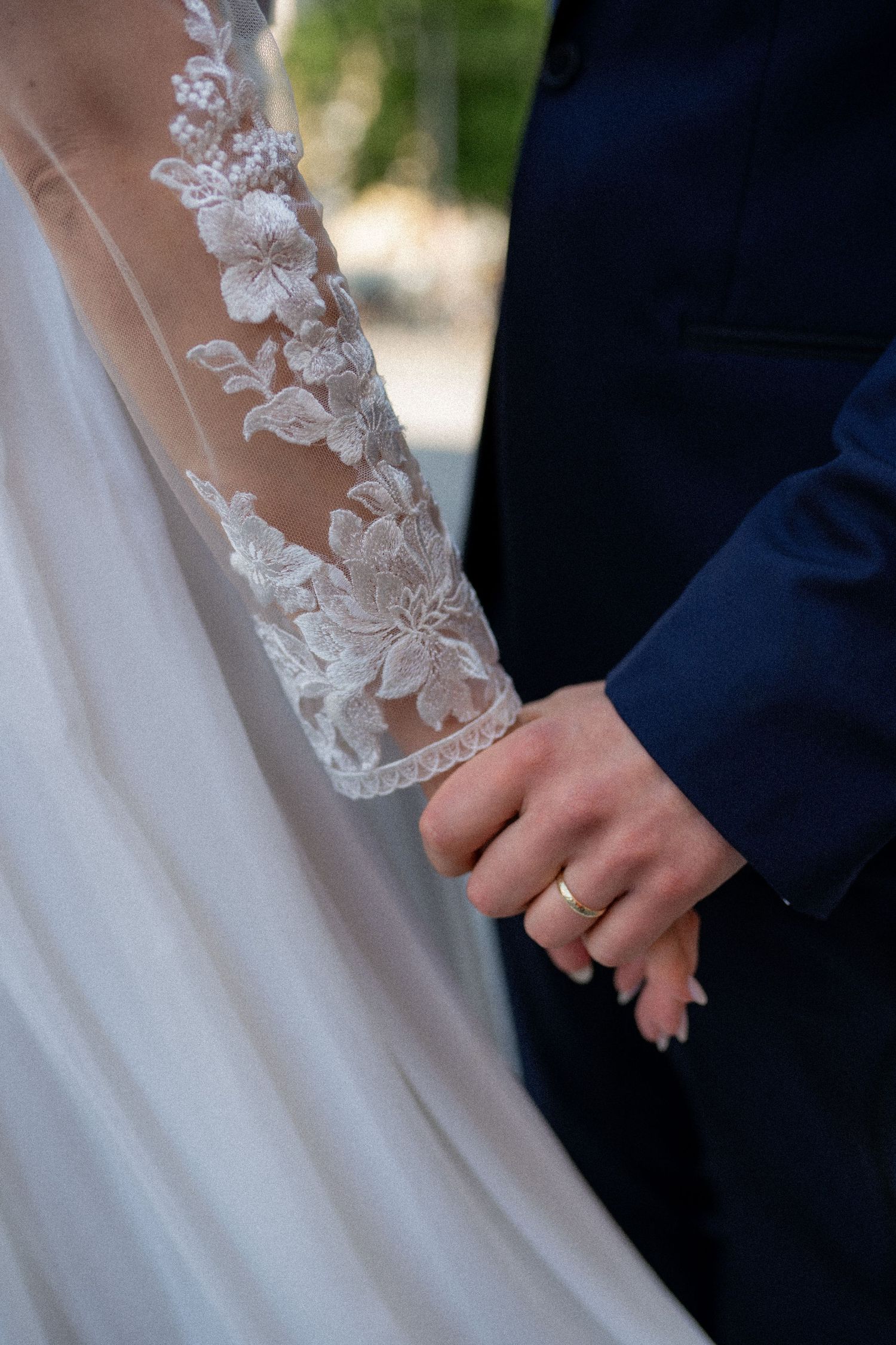 Close-up of bride and groom holding hands showing wedding ring, bride wearing elegant lace sleeve wedding dress.