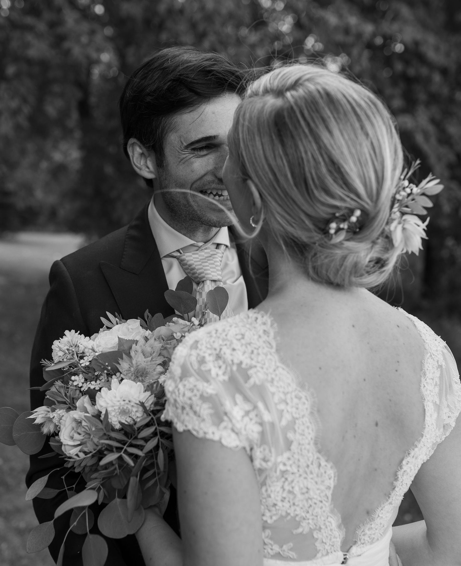 Black and white photo of smiling groom and bride holding a floral bouquet outdoors.