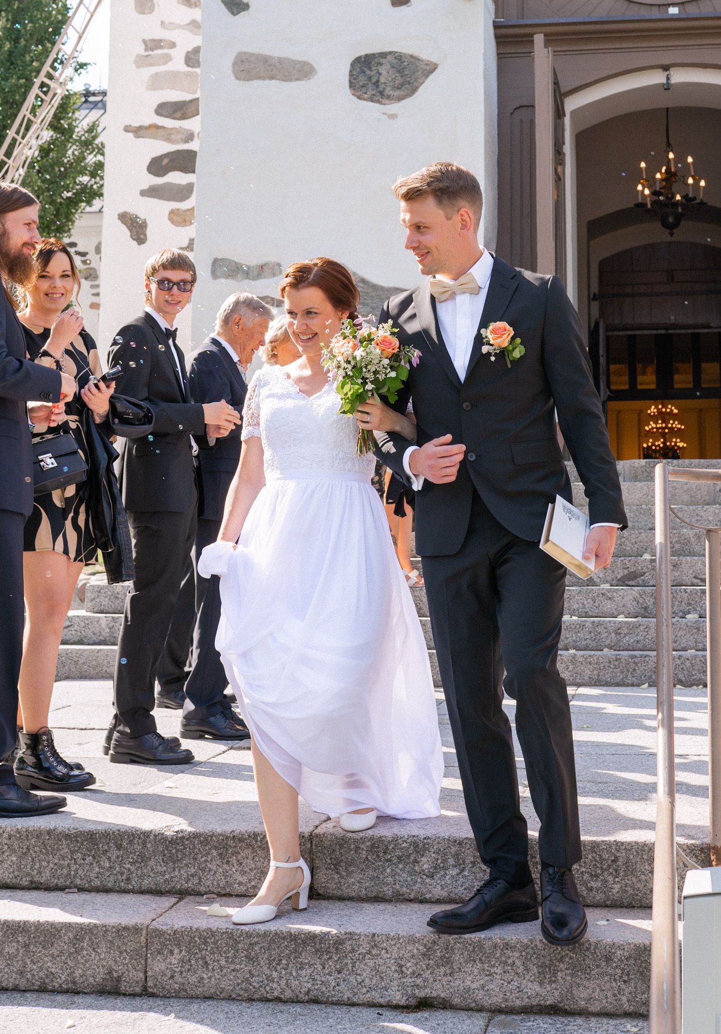 Bride holding bouquet and groom in black suit walk down church steps on wedding day.