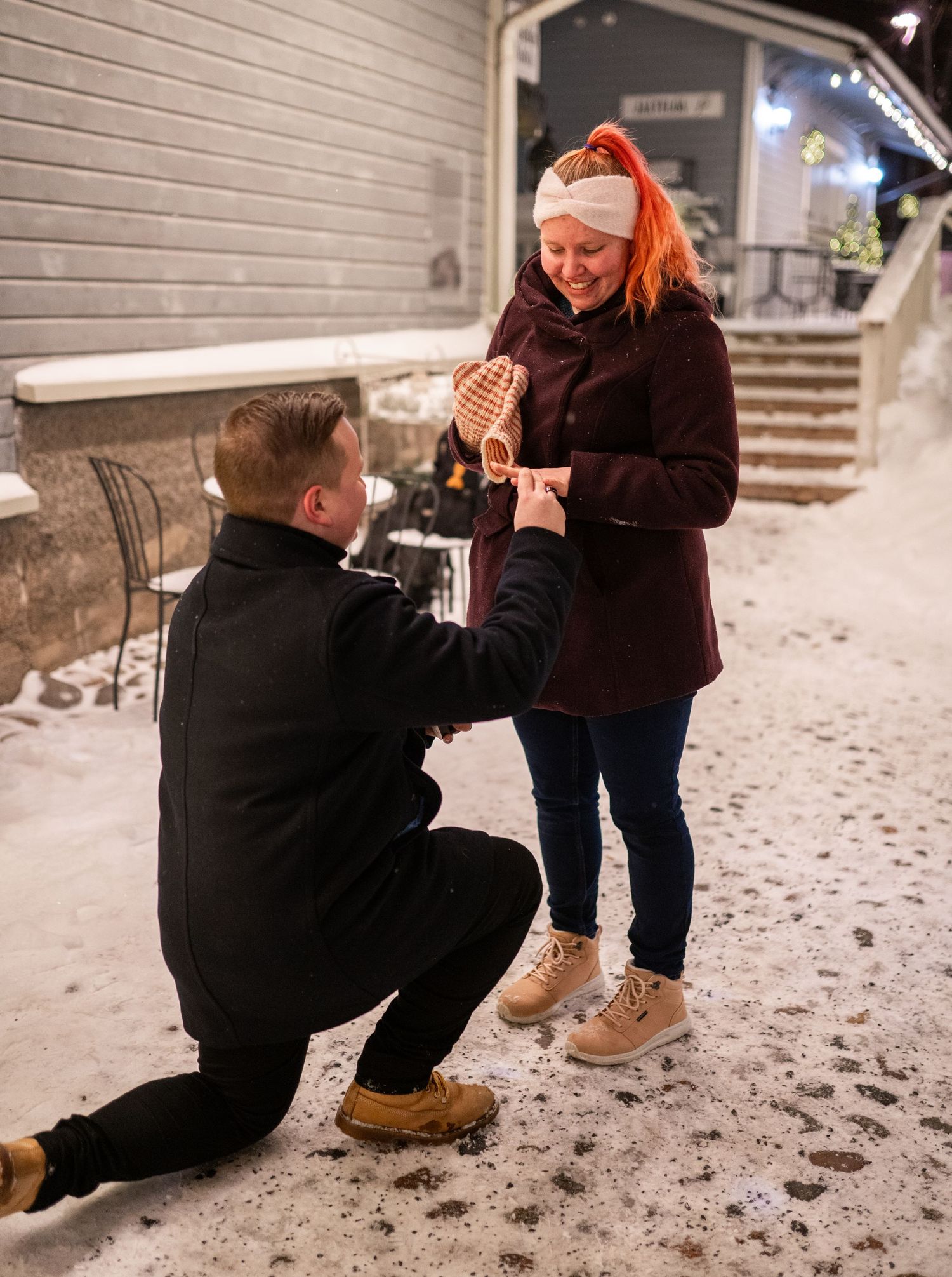 Man kneeling on snowy ground proposing to red-haired woman in dark coat outdoors at night.