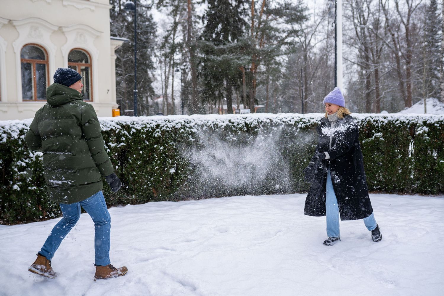 Two people having a snowball fight outdoors in winter, wearing colorful coats and hats near snow-covered hedges.