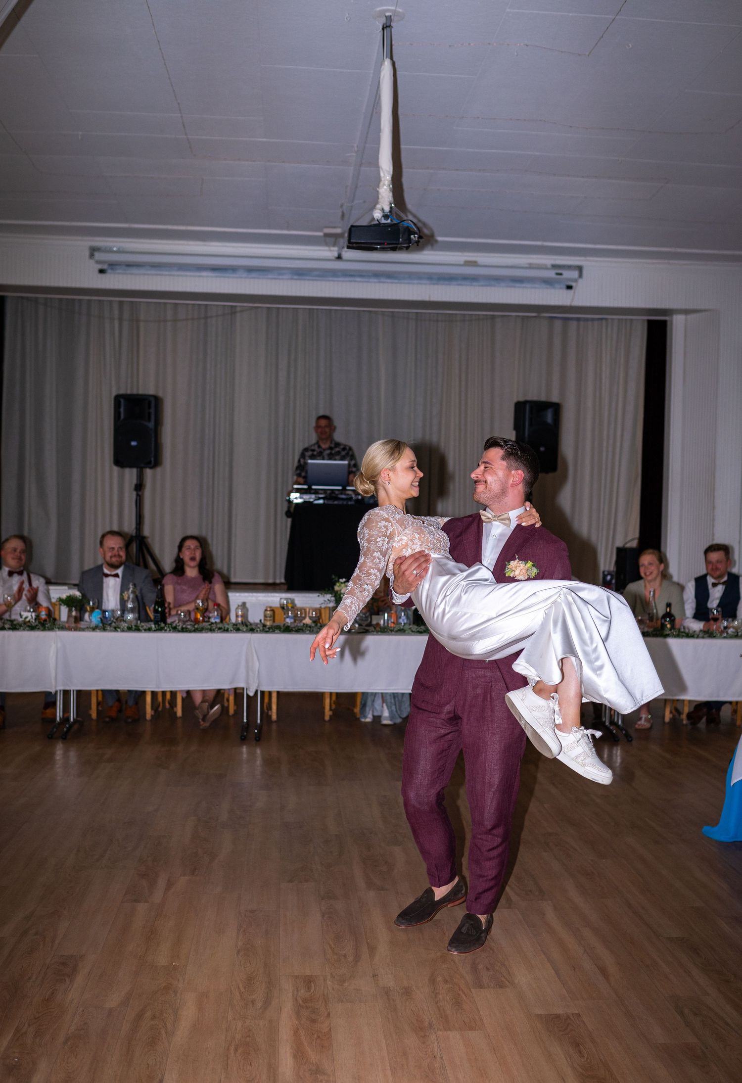 Groom carries smiling bride in his arms during first dance at wedding reception with guests watching.