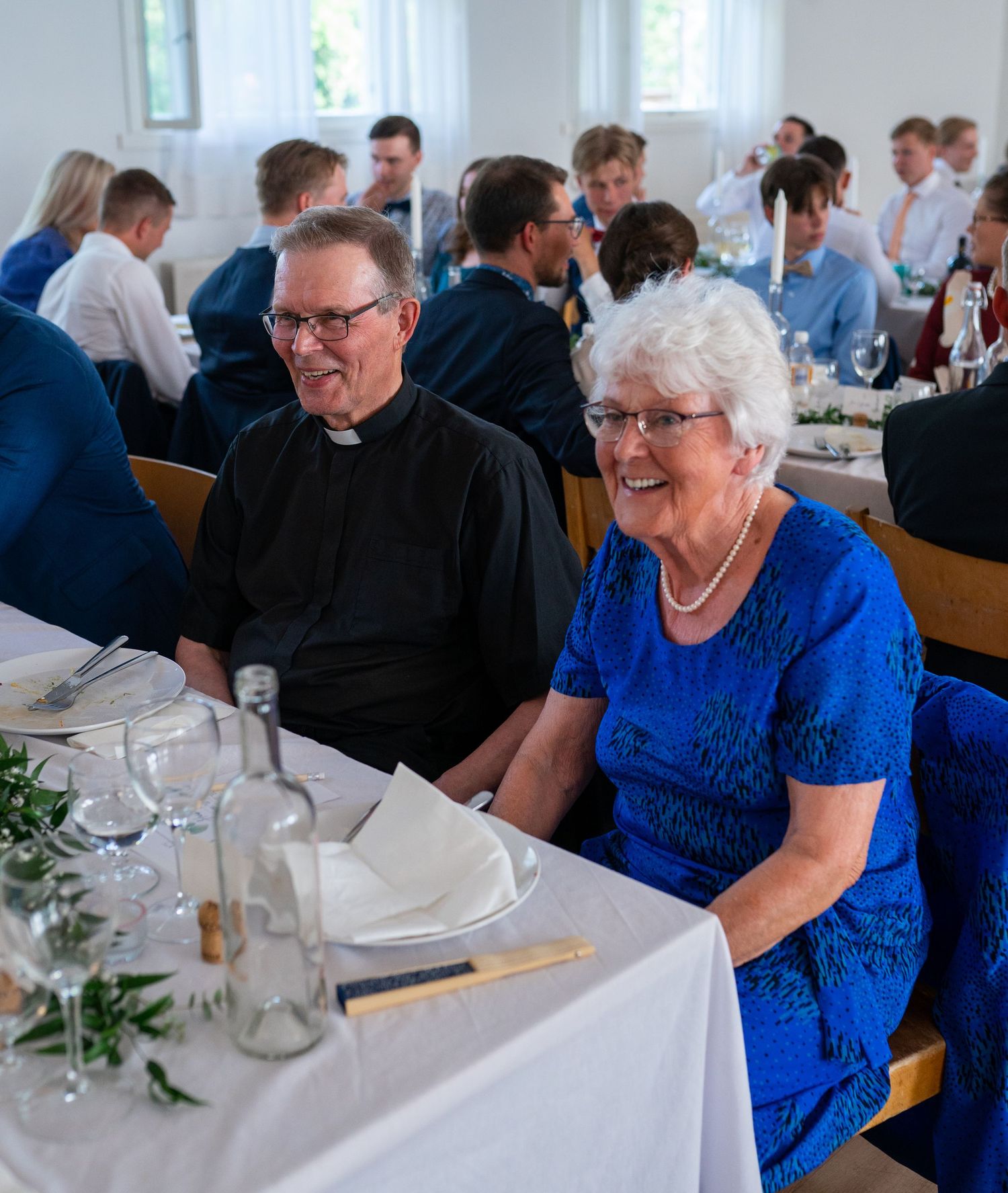 Elderly couple smiling warmly at a wedding reception, man wearing a clerical collar.