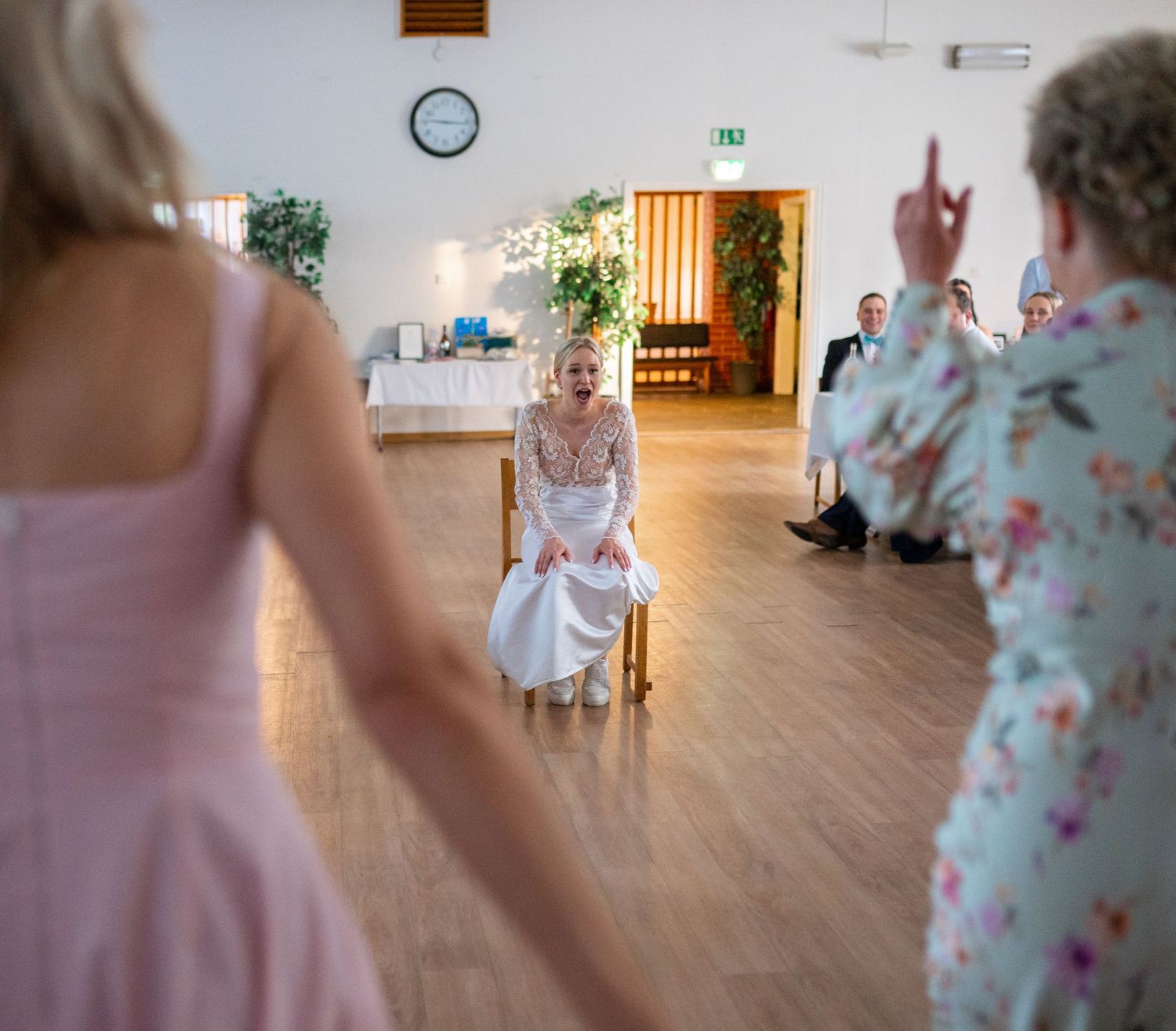 Bride in white lace dress seated on chair laughing as guests perform surprise wedding dance.