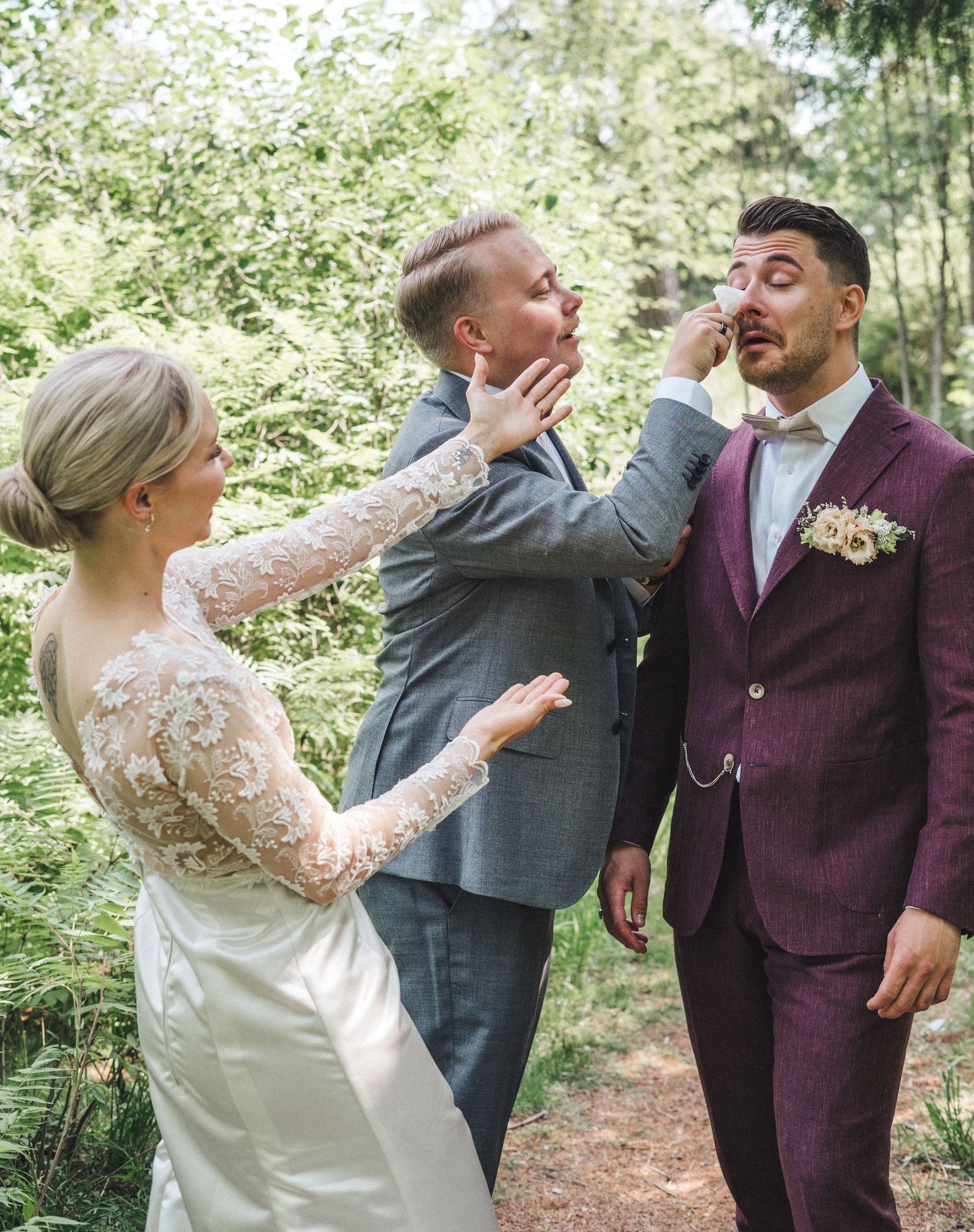 Groomsman in gray suit playfully wipes tears from emotional groom in burgundy suit as bride watches.