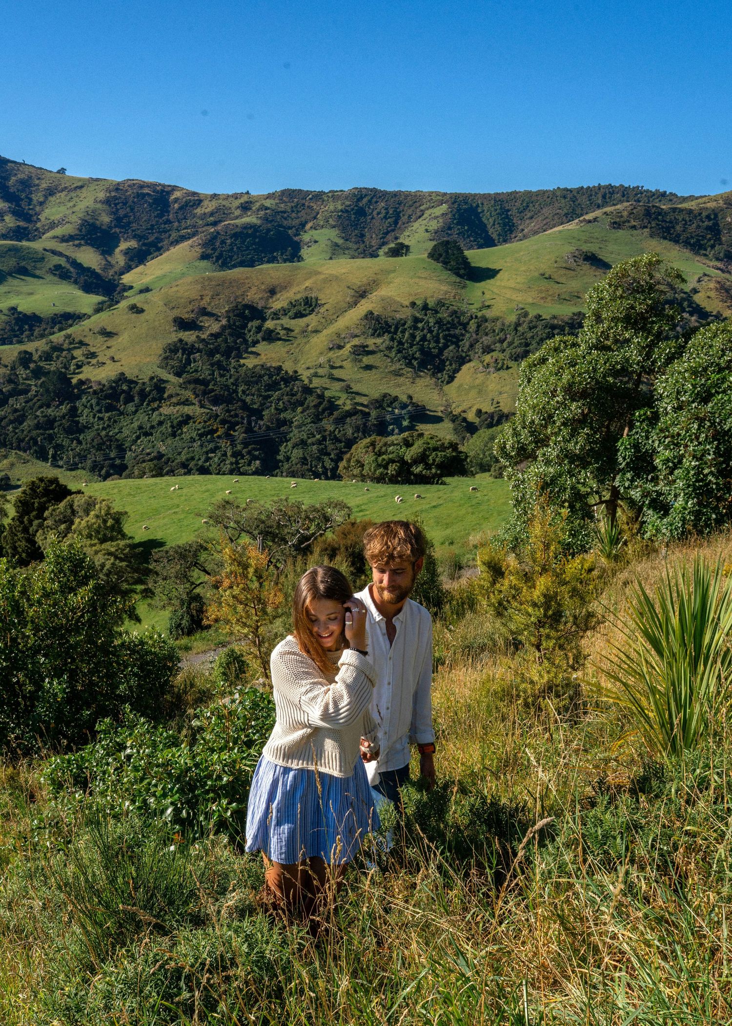 A couple explores lush green hillside vegetation with sheep grazing in the valley and mountains beyond.