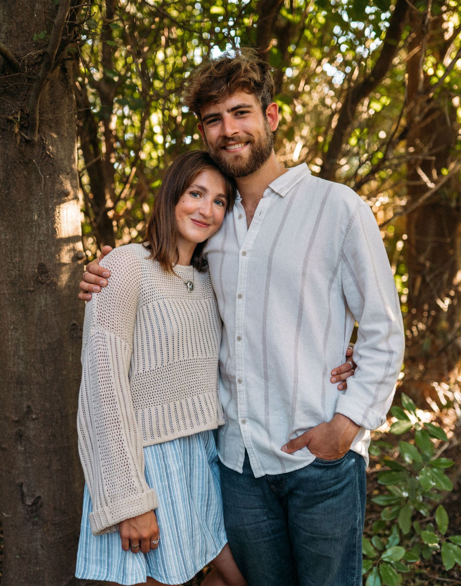 Couple posing together by a tree, woman in knit sweater and man in white shirt.