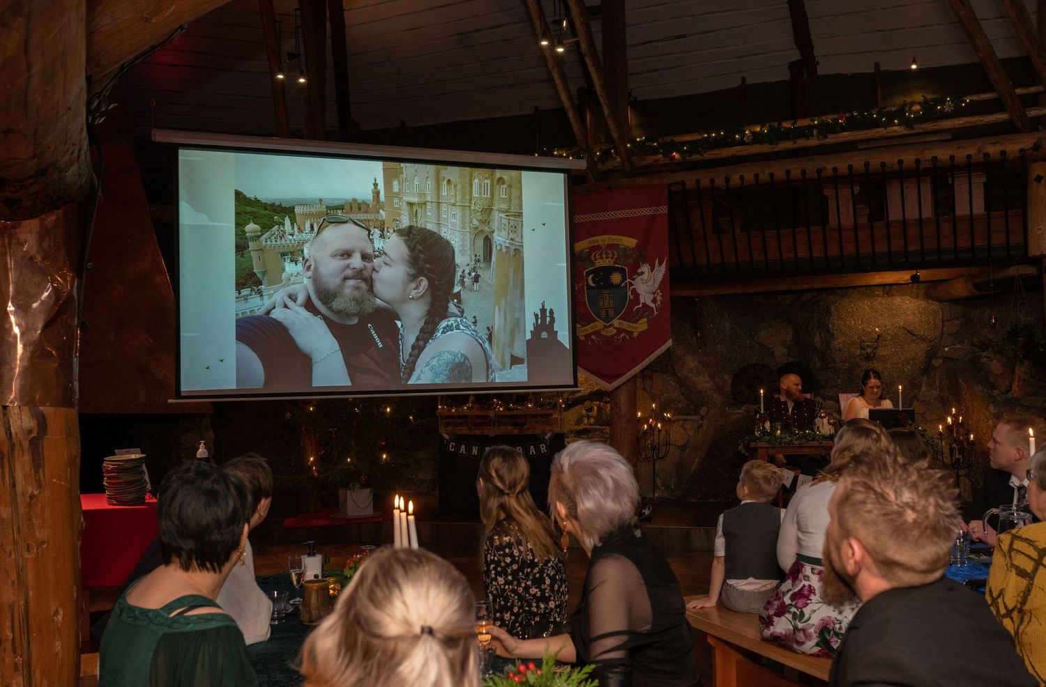 Guests watch a couple's photo on a projector screen inside a rustic medieval-themed venue during a celebration.