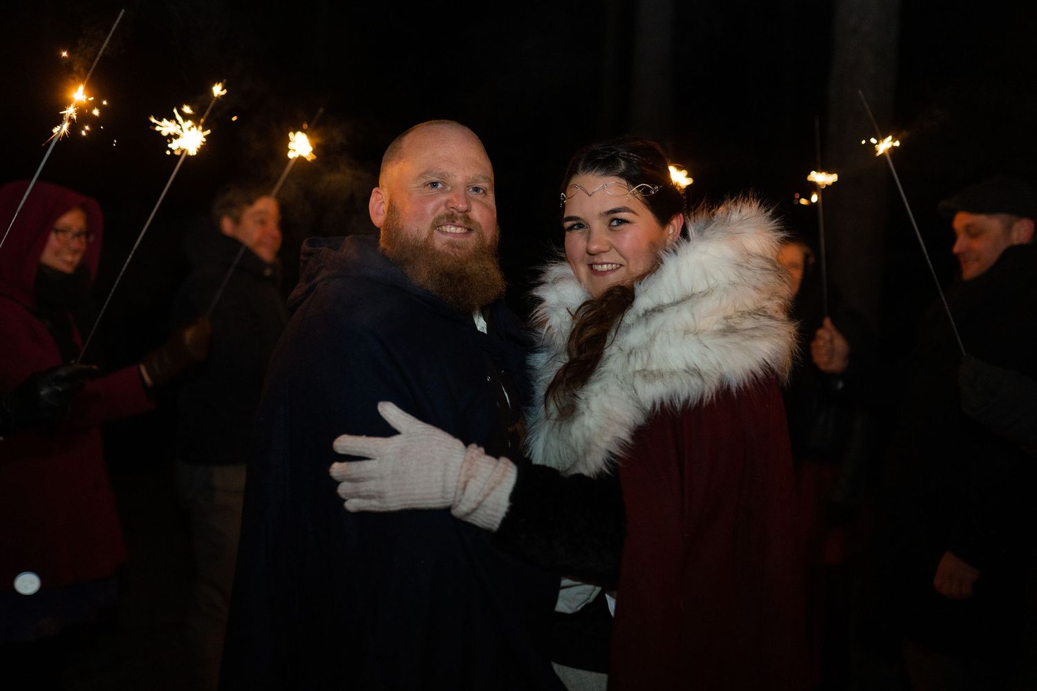 Couple smiling and embracing outdoors at night, surrounded by people holding sparklers at a celebration.