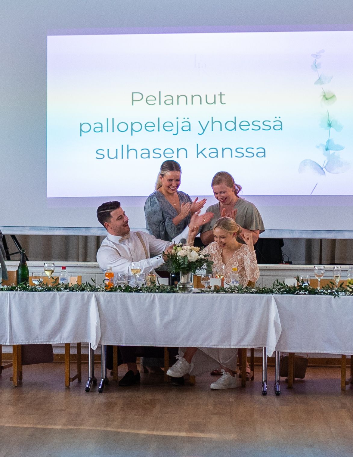 Wedding reception toast scene with bride and groom at head table, Finnish text displayed on screen behind.