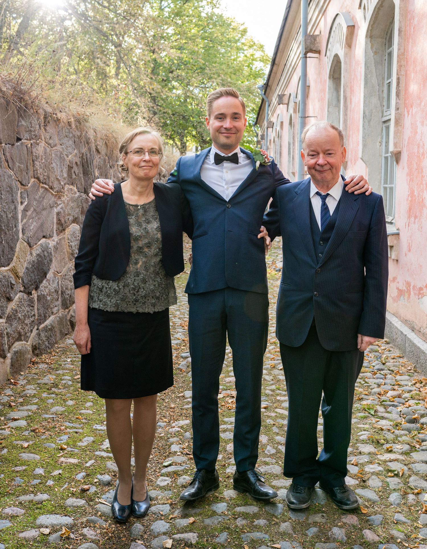 Groom in navy tuxedo with bow tie poses between elderly parents on cobblestone path by stone wall.