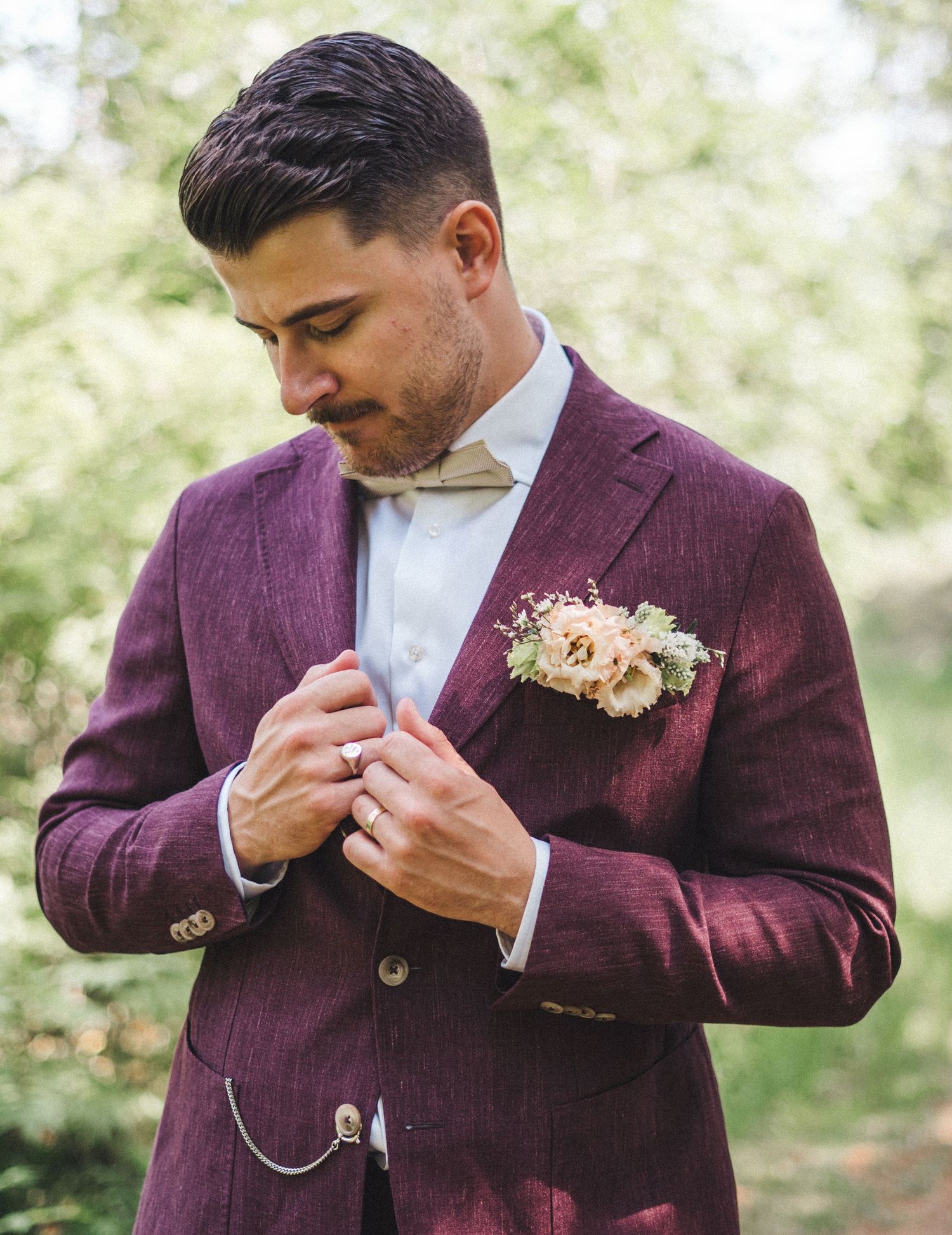 Groom in deep burgundy linen suit adjusting jacket with floral boutonniere outdoors.