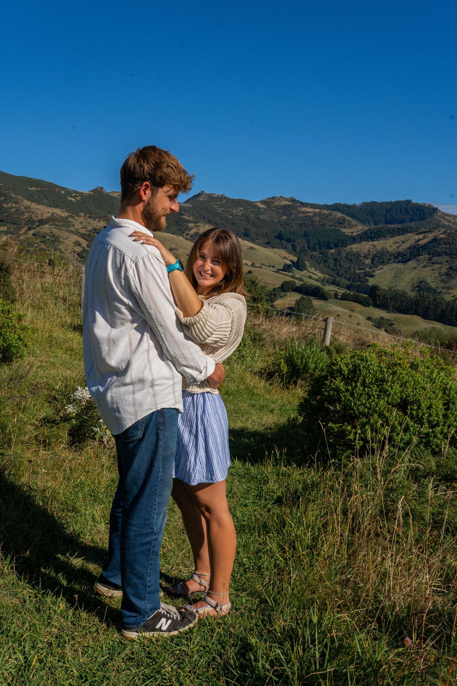 Smiling couple hugs closely on sunlit green hillside with scenic mountain range behind.