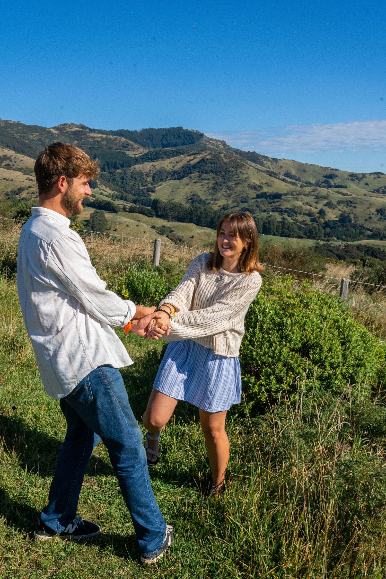 Laughing couple holds hands playfully on sunny green hillside with mountain valley views.