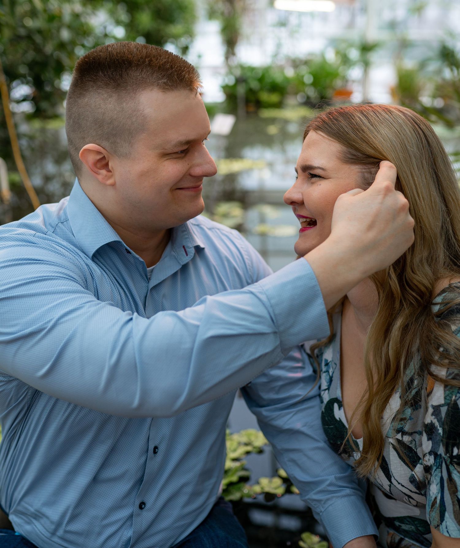 A couple shares a tender moment in a lush greenhouse, man gently touching womans face as she smiles joyfully.