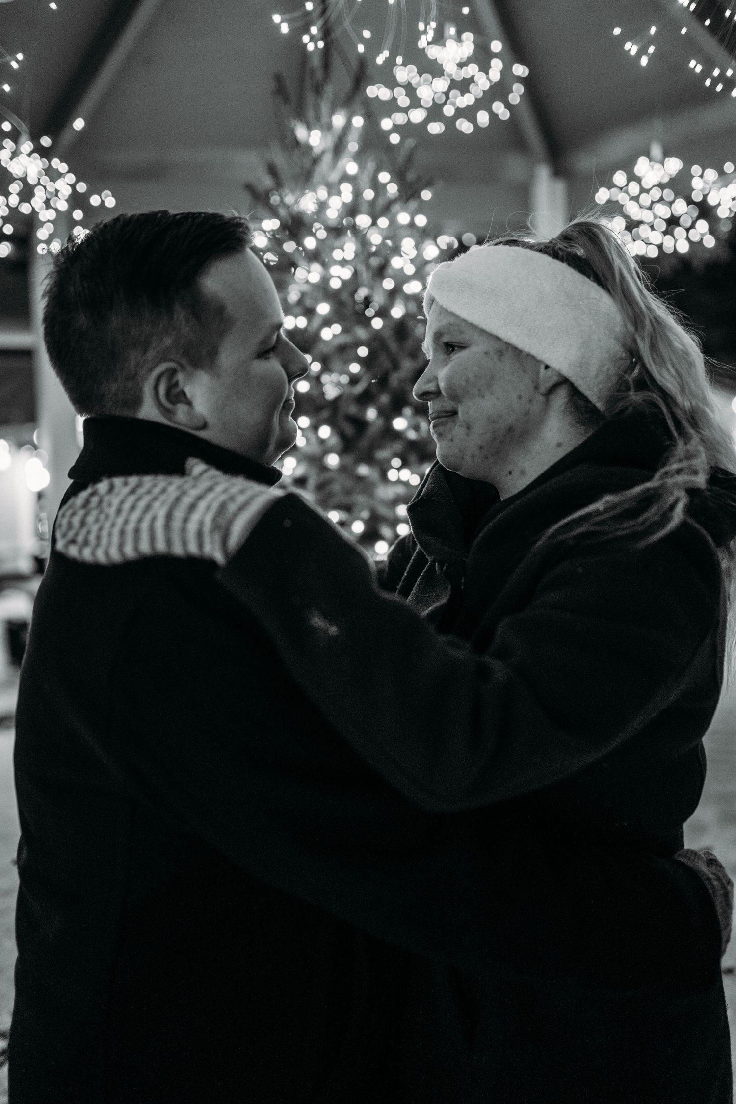 Black and white photo of a couple embracing near a lit Christmas tree at night.