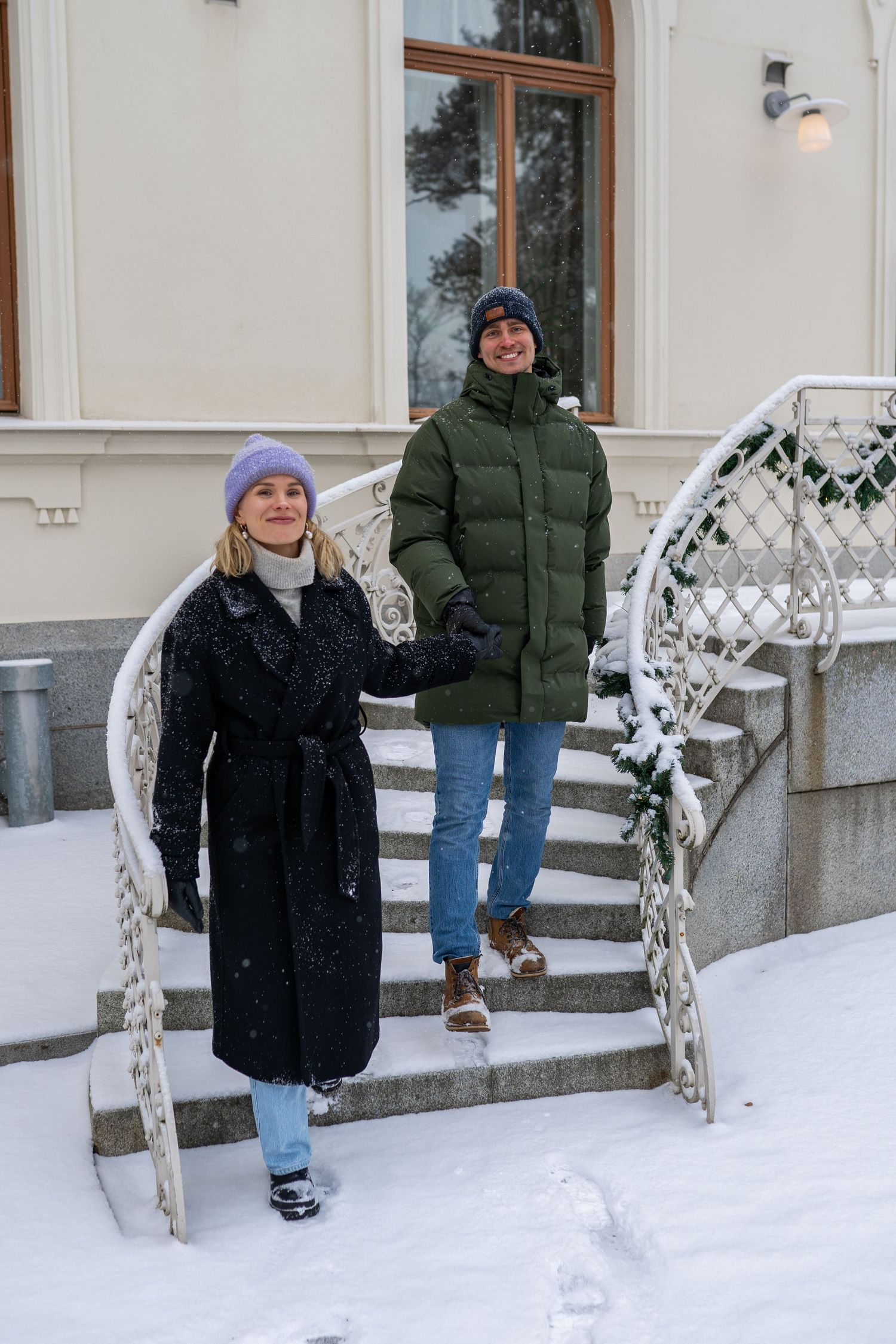 Couple in winter coats descending snow-covered stairs outside a classic building on a snowy day.