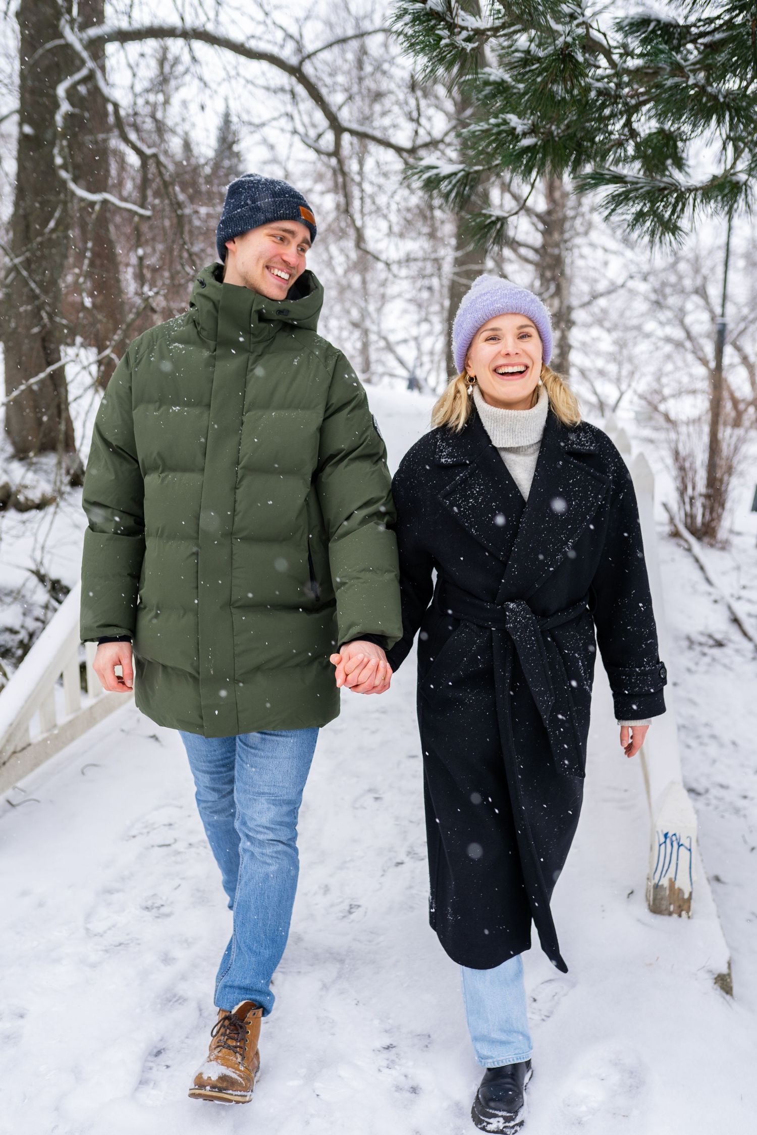 Happy couple holding hands walking through snowy winter park, wearing warm coats and hats during snowfall.