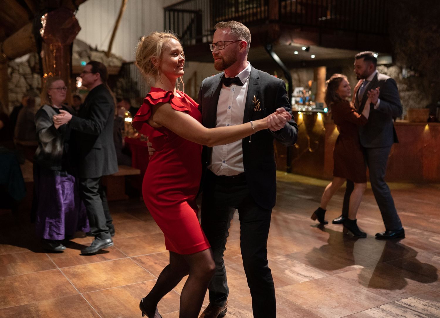 Couple dancing elegantly at a formal event, woman in red dress, man in tuxedo, guests socializing in background.