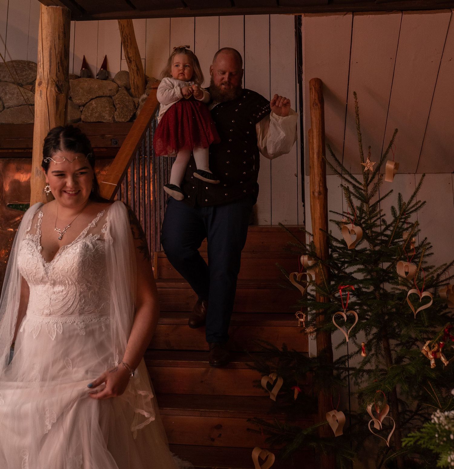 Bride in white lace gown stands before groom carrying child in red dress down rustic wooden stairs near Christmas tree.