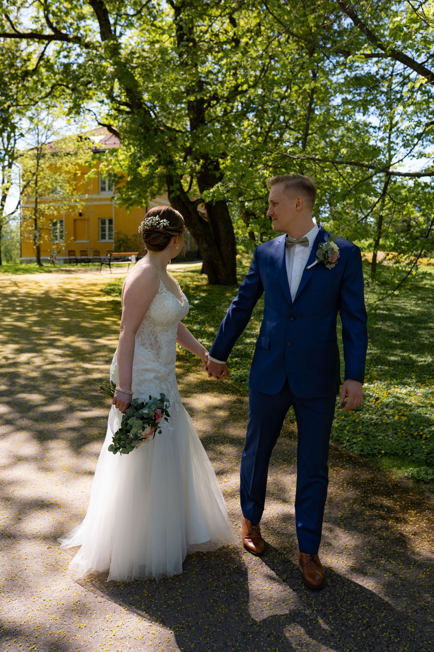 Bride and groom walk hand in hand through sunny park with large trees and yellow building behind.