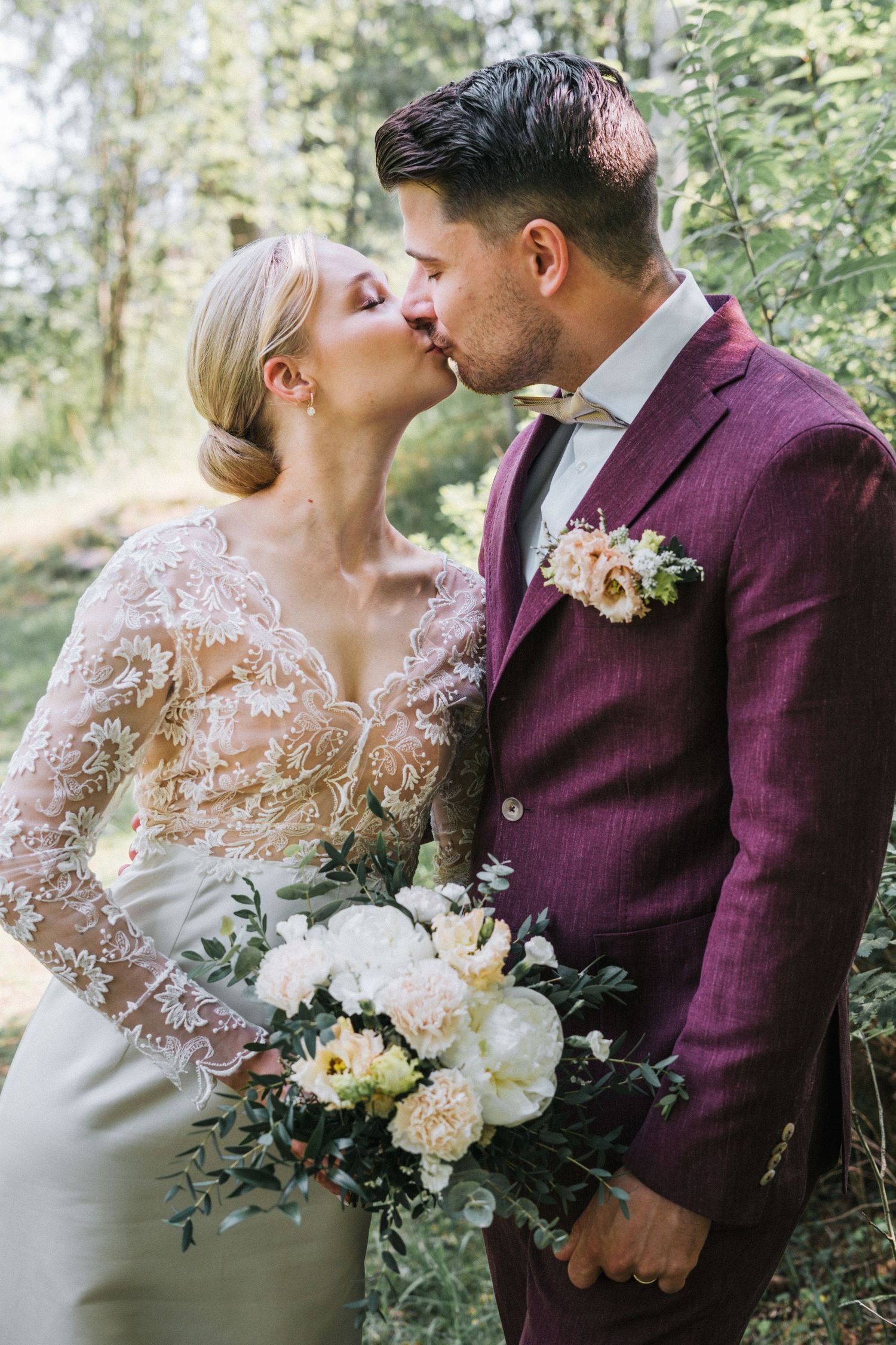 Bride and groom sharing a romantic kiss outdoors, bride holding white peony bouquet, groom in burgundy suit.