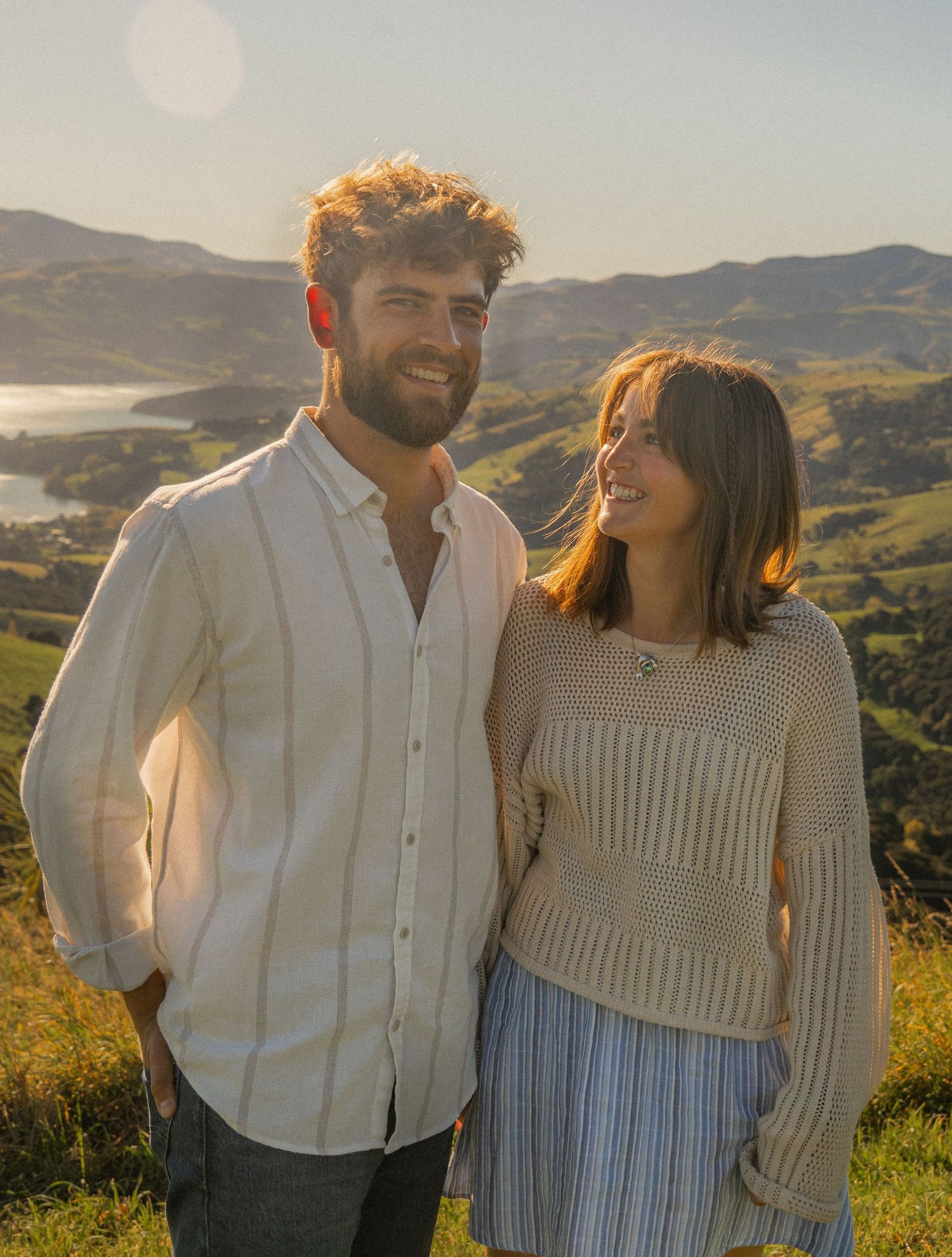 Smiling couple posing together on a hilltop with a stunning coastal bay backdrop at sunset.
