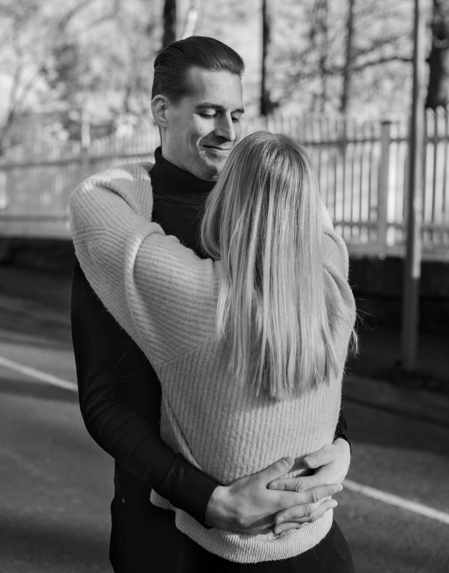 Black and white photo of a smiling man hugging a blonde woman outdoors near a fence.